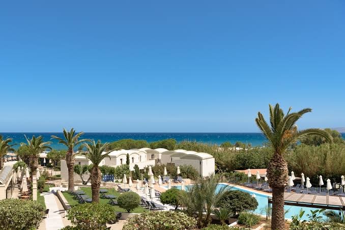 Agapi Beach Resort panorāmas skats uz Vidusjūru, viesnīcas ēkām, palmām un baseinu zonu saulainā dienā
