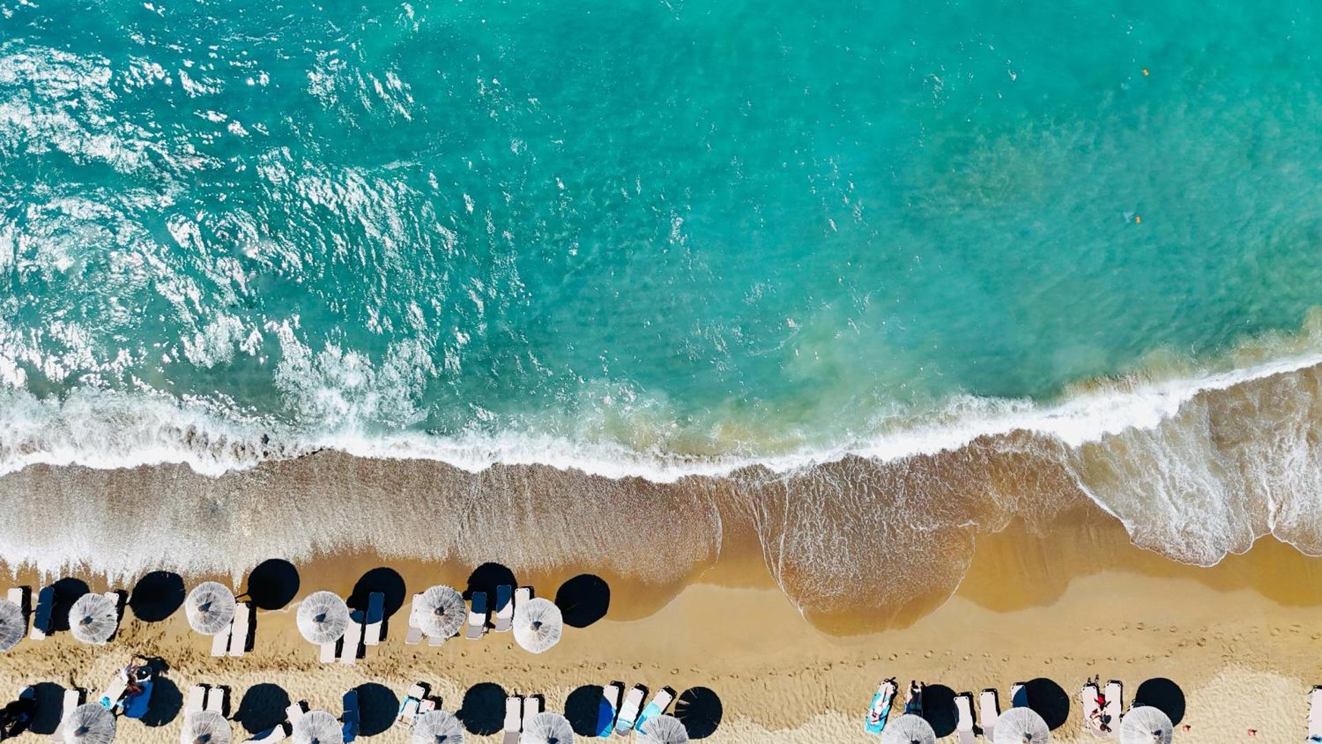 Agapi Beach Resort tirkīza krāsas jūras ūdens un gaišas smiltis ar saulessargu rindām no augšas