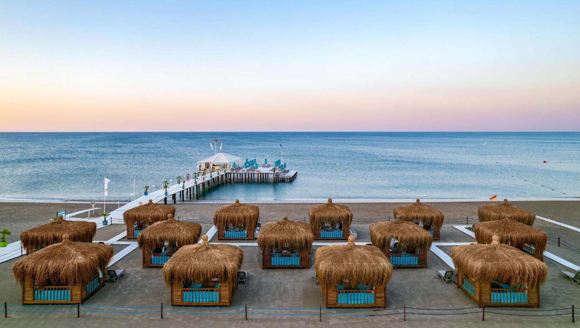 Concorde De Luxe Resort ekskluzīvas atpūtas nojumes jeb bungalo pludmales smiltīs saulrieta laikā ar skatu uz jūras piestātni