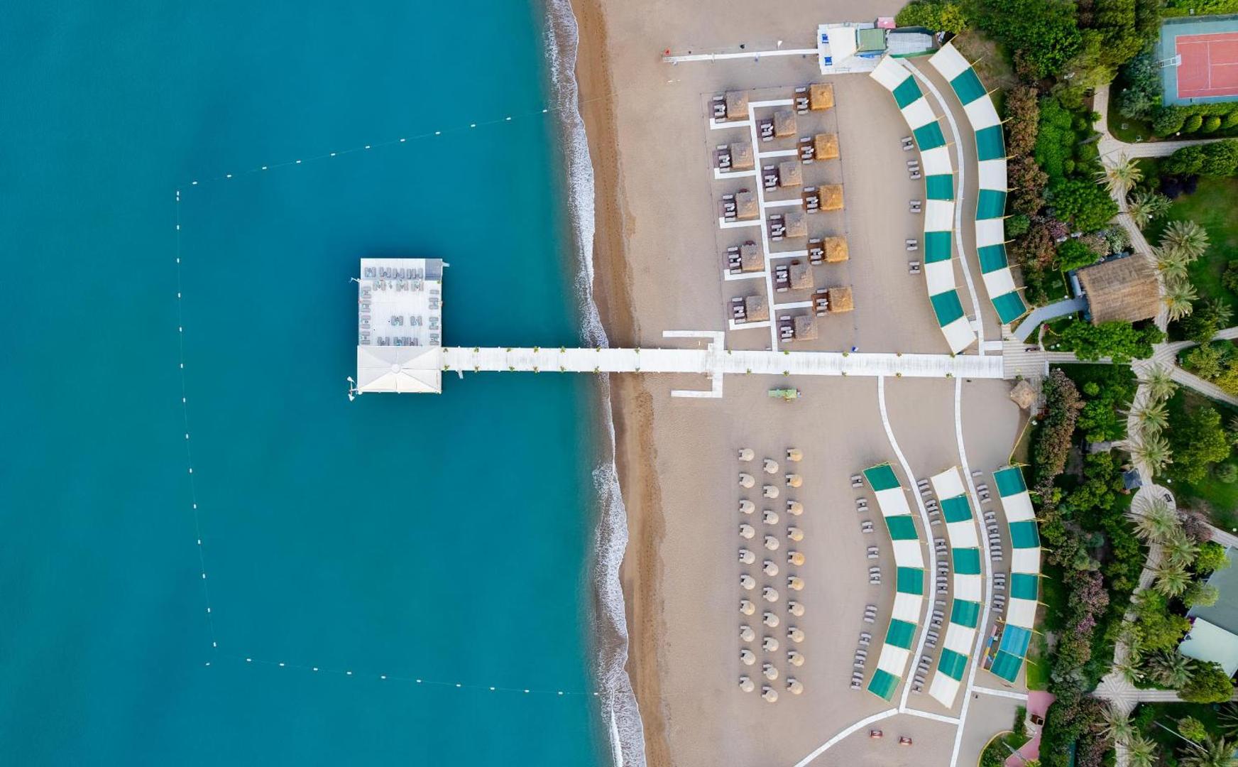 Concorde De Luxe Resort privātā pludmale no augšas ar piestātni, saulessargiem un tirkīzzilu jūras ūdeni Antaljas kūrortā.