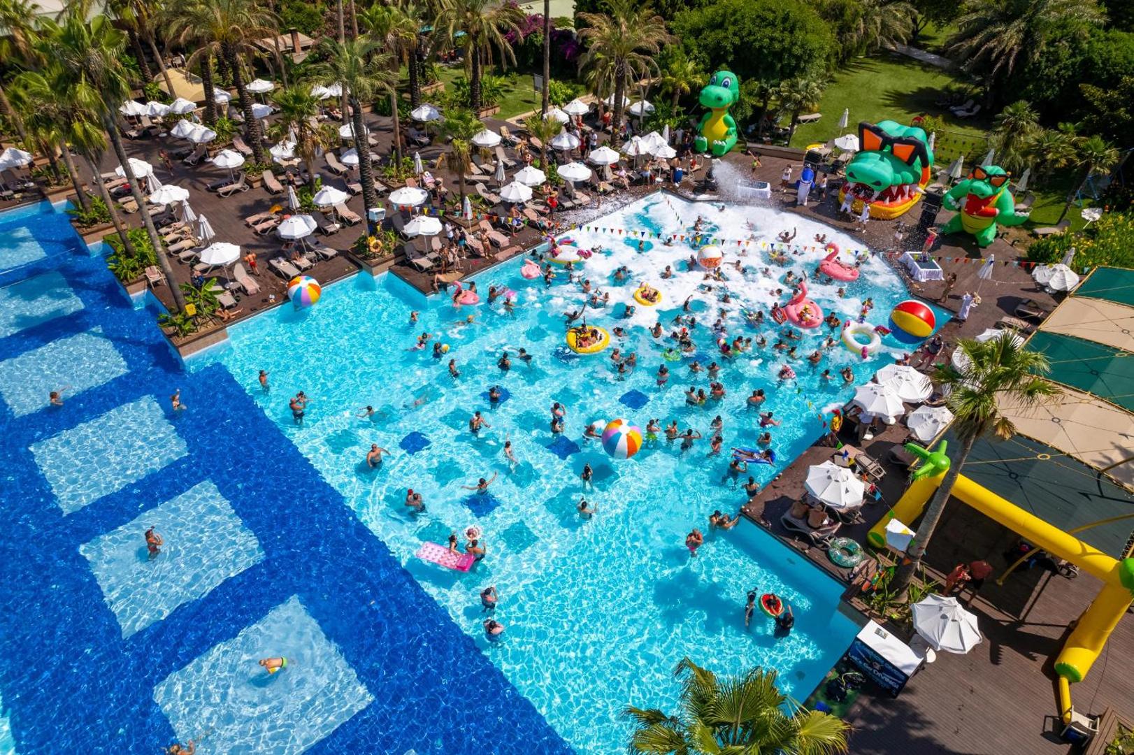 Aktīva atpūta Concorde De Luxe Resort galvenajā baseinā ar piepūšamām atrakcijām, palmām un atpūtas krēsliem saulainā dienā.