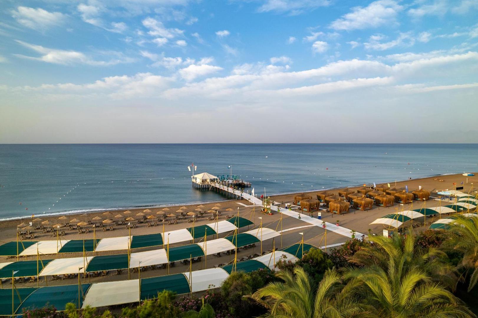 Concorde De Luxe Resort plašā smilšu pludmale Antaljā ar saulessargiem, nojumēm un garu koka piestātni Vidusjūras piekrastē