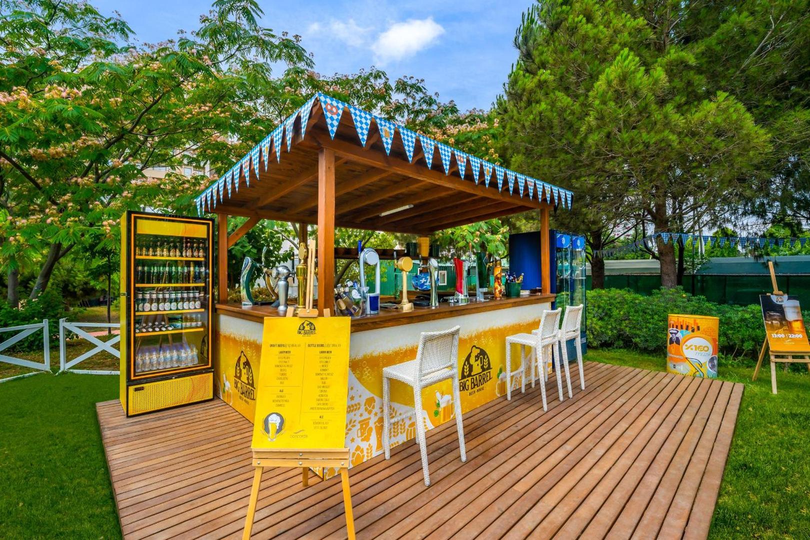 Concorde De Luxe Resort āra bārs Big Barrel dārzā ar koka terasi, bāra krēsliem un alus dzesētājiem dienas gaismā