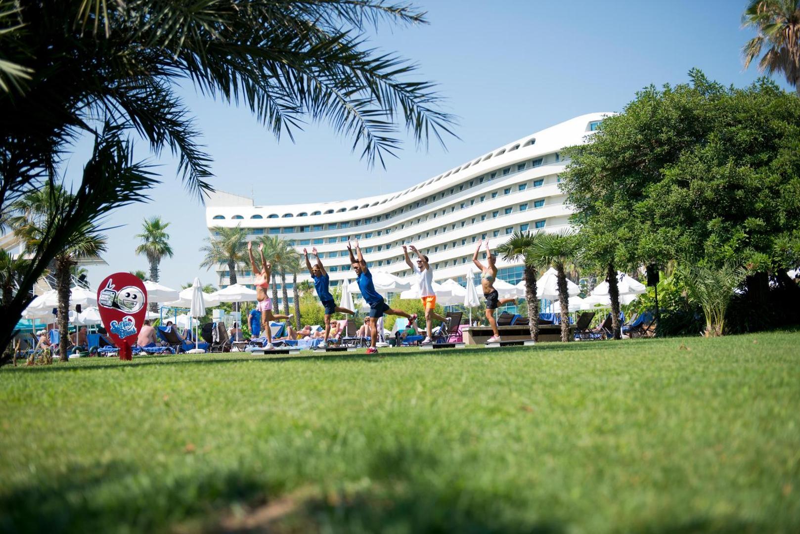 Viesnīcas Concorde De Luxe Resort galvenā ēka un rīta vingrošana uz zaļā zālāja palmu ieskautā dārzā Larā