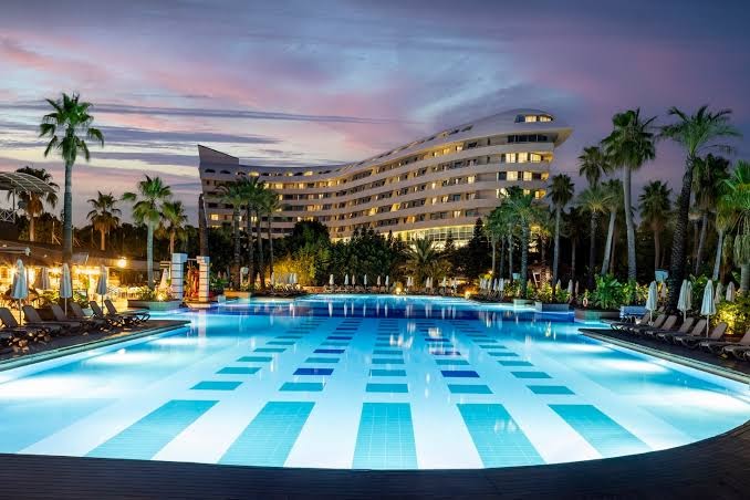 Vakara skats uz Concorde De Luxe Resort moderno arhitektūru, izgaismotu baseinu un palmām saulrieta laikā Antaljā.