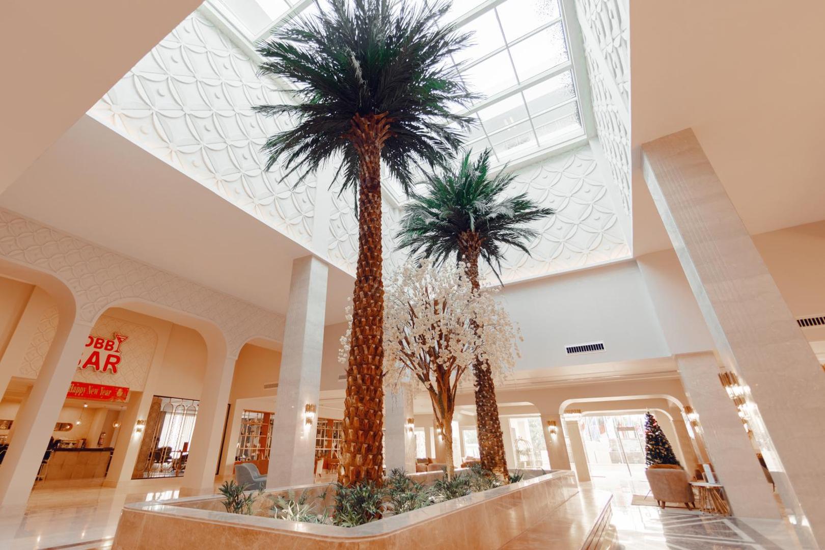 Bellagio Beach Resort & Spa viesnīcas vestibils ar augstām palmām un stikla griestiem — gaiša un moderna koplietošanas telpa