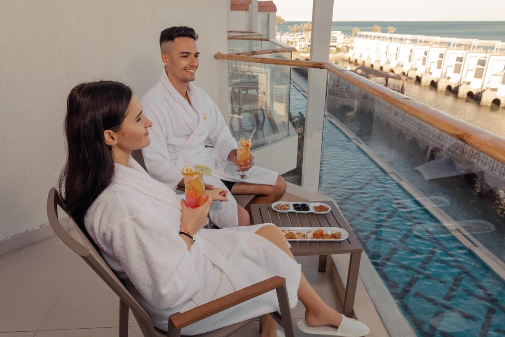 Pāris baltos halātos bauda kokteiļus uz Bellagio Beach Resort & Spa balkona ar skatu uz baseinu un jūru