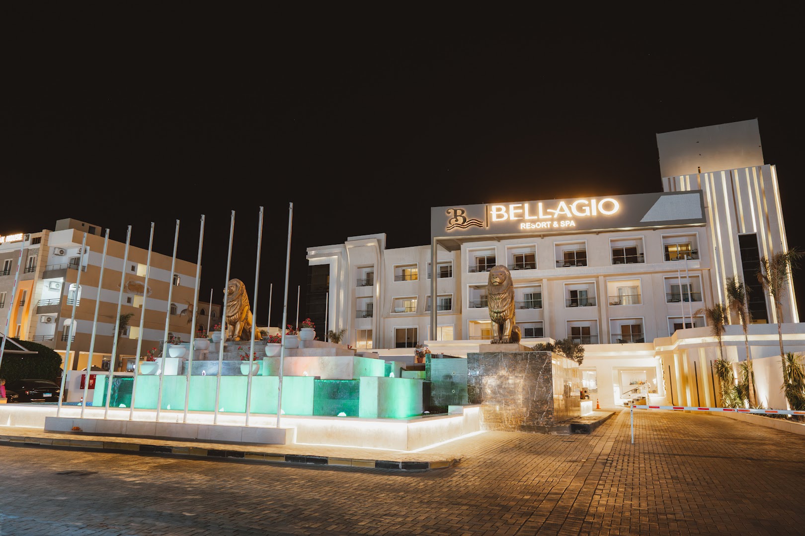 Bellagio Beach Resort & Spa galvenā ieeja naktī ar izgaismotu strūklaku, lauvu skulptūrām un viesnīcas logo Hurgadā, Ēģiptē