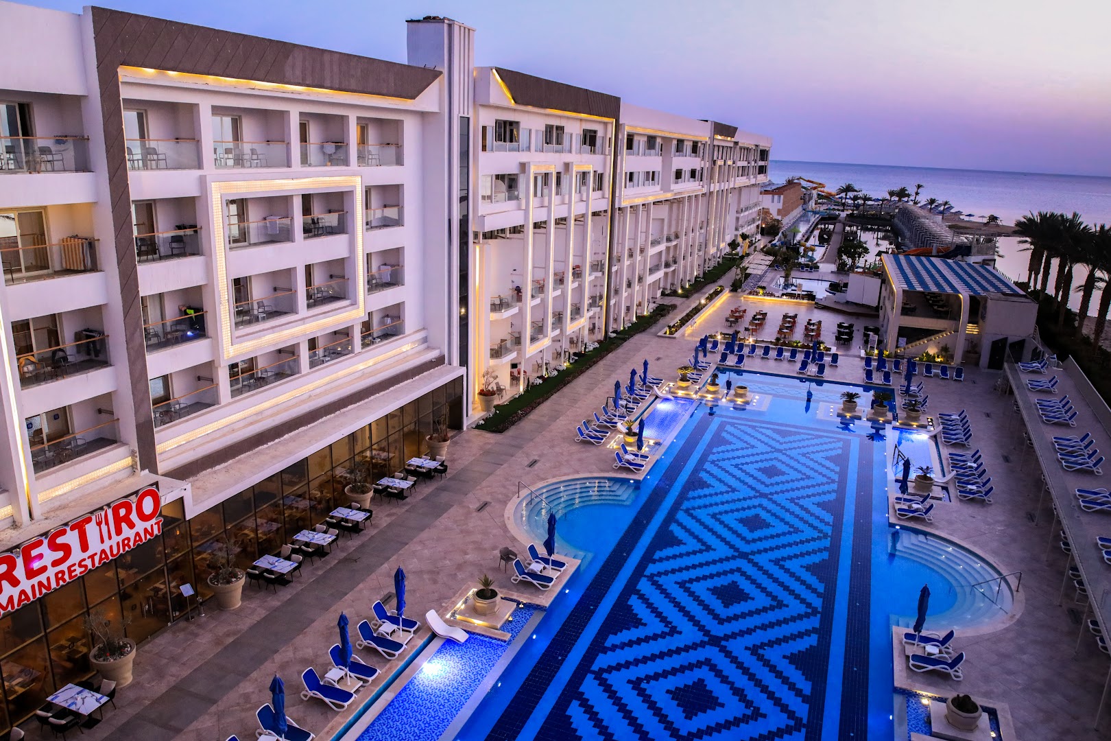 Bellagio Beach Resort & Spa Hurgadā skats no augšas uz galveno baseinu ar rakstainu flīžu klājumu, restorāna terasi un viesnīcas korpusu vakarā