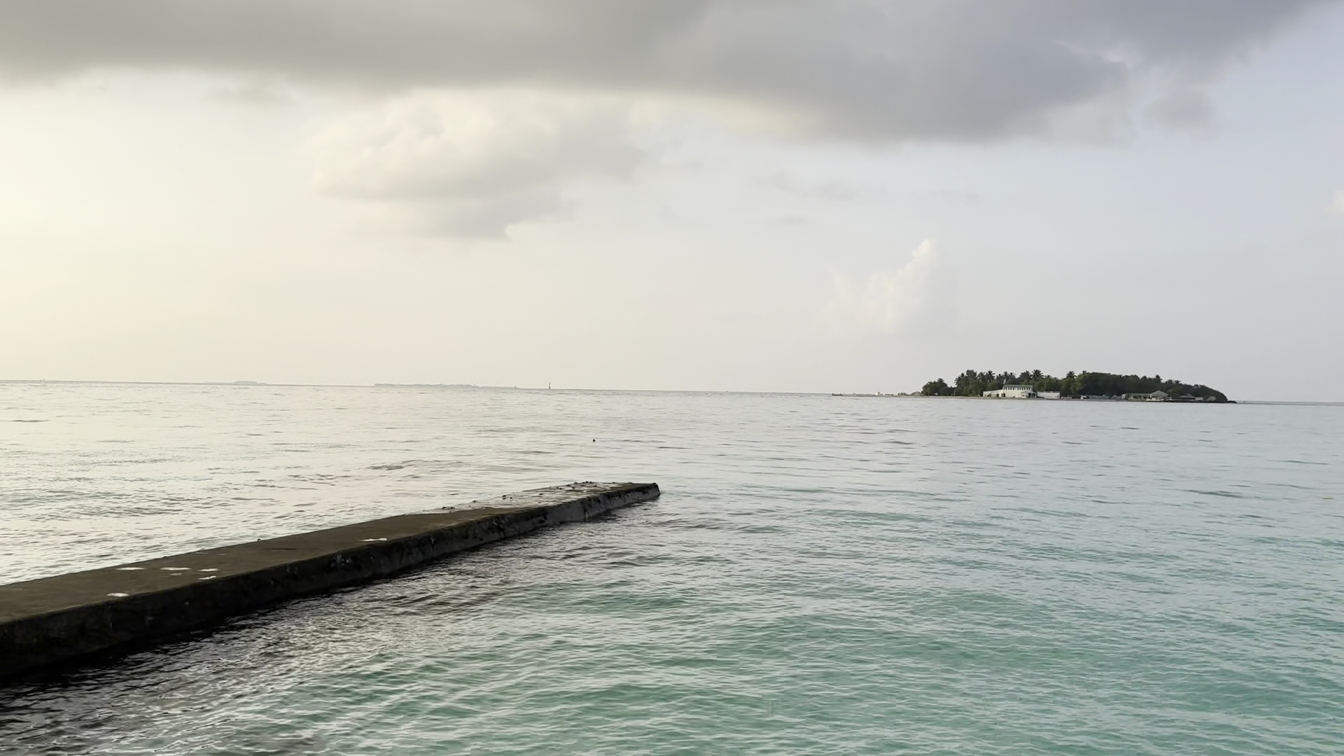Sheraton Maldives Full Moon Resort & Spa skats no mola uz mierīgu okeānu un kaimiņu salu mākoņainā dienā
