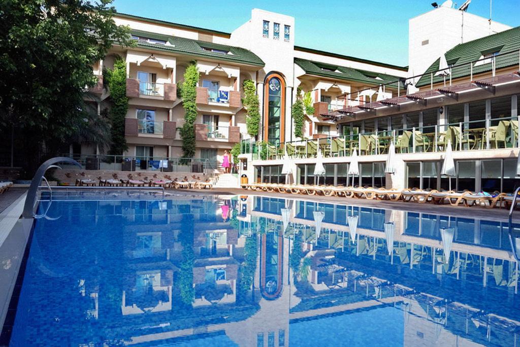 Ambassador Plaza Hotel baseina zona ar ēkas atspulgu ūdenī, terasi un zaļojošiem augiem pie fasādes Turcijā