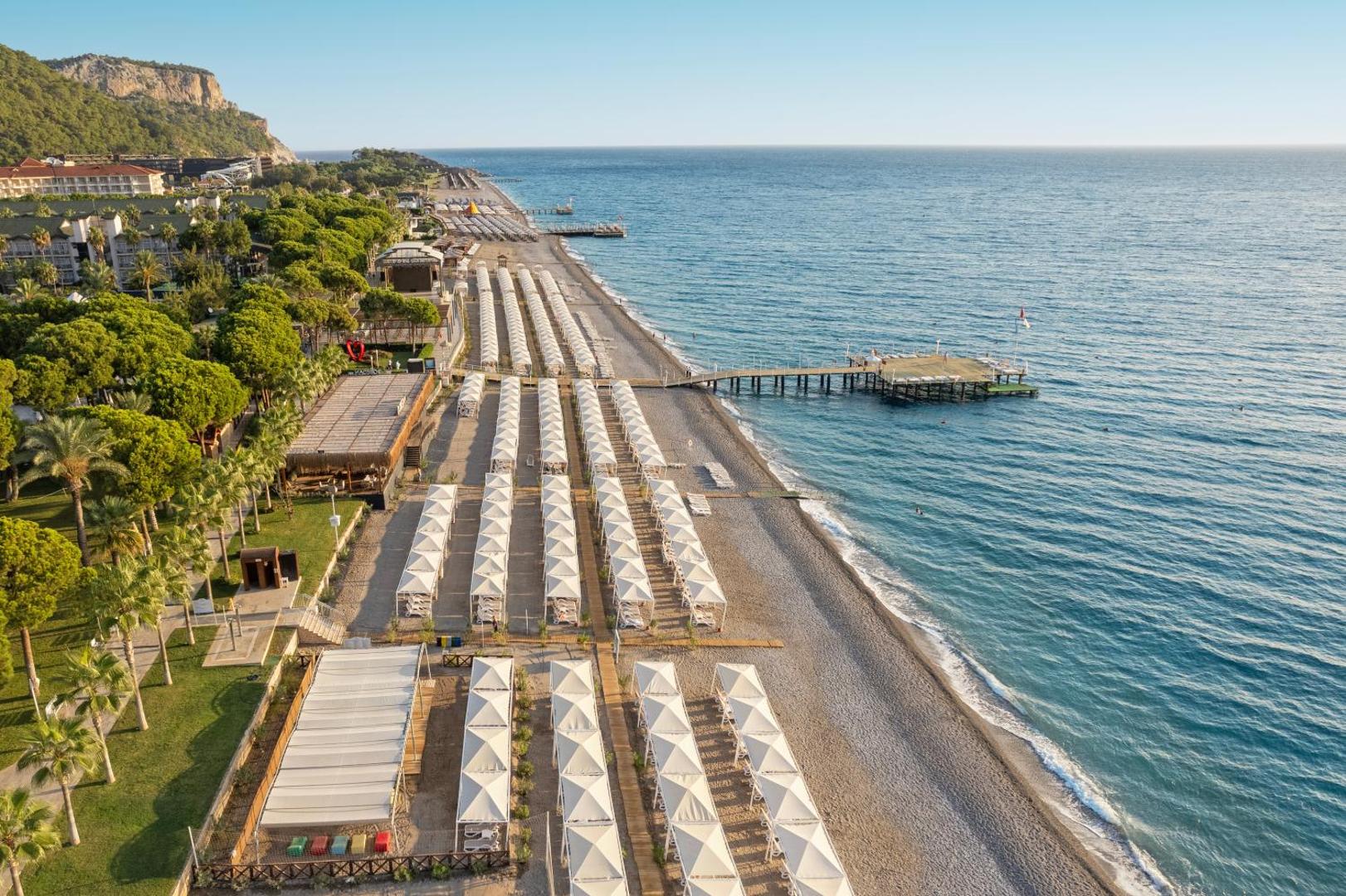 Dobedan World Palace plašā pludmales līnija ar saulessargiem, koka laipu un piestātni Vidusjūras piekrastē, Turcijā