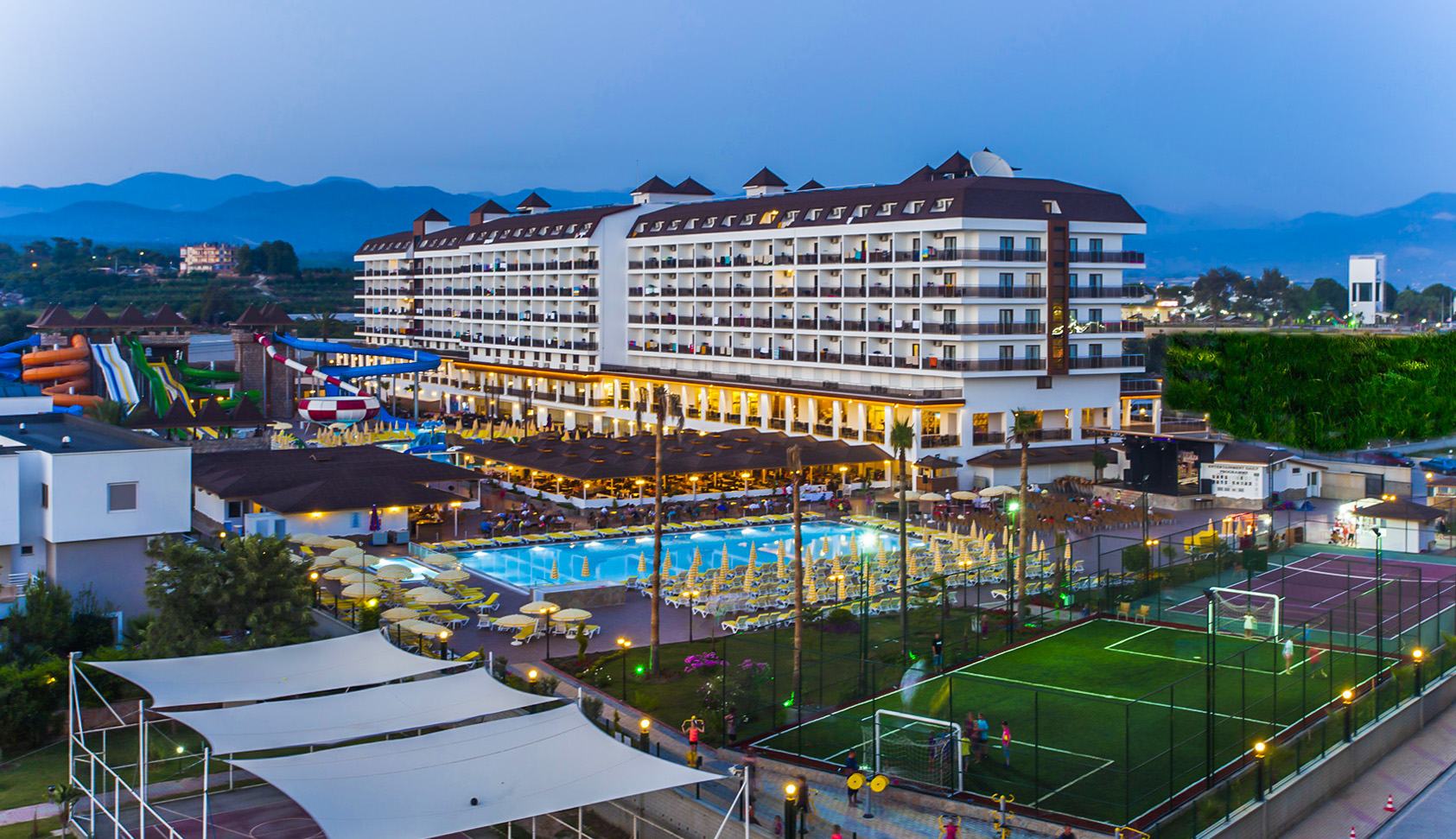 Eftalia Splash Resort vakara apgaismojumā — galvenā ēka, izgaismots āra baseins, futbola laukums un tenisa korts Alanjas reģionā