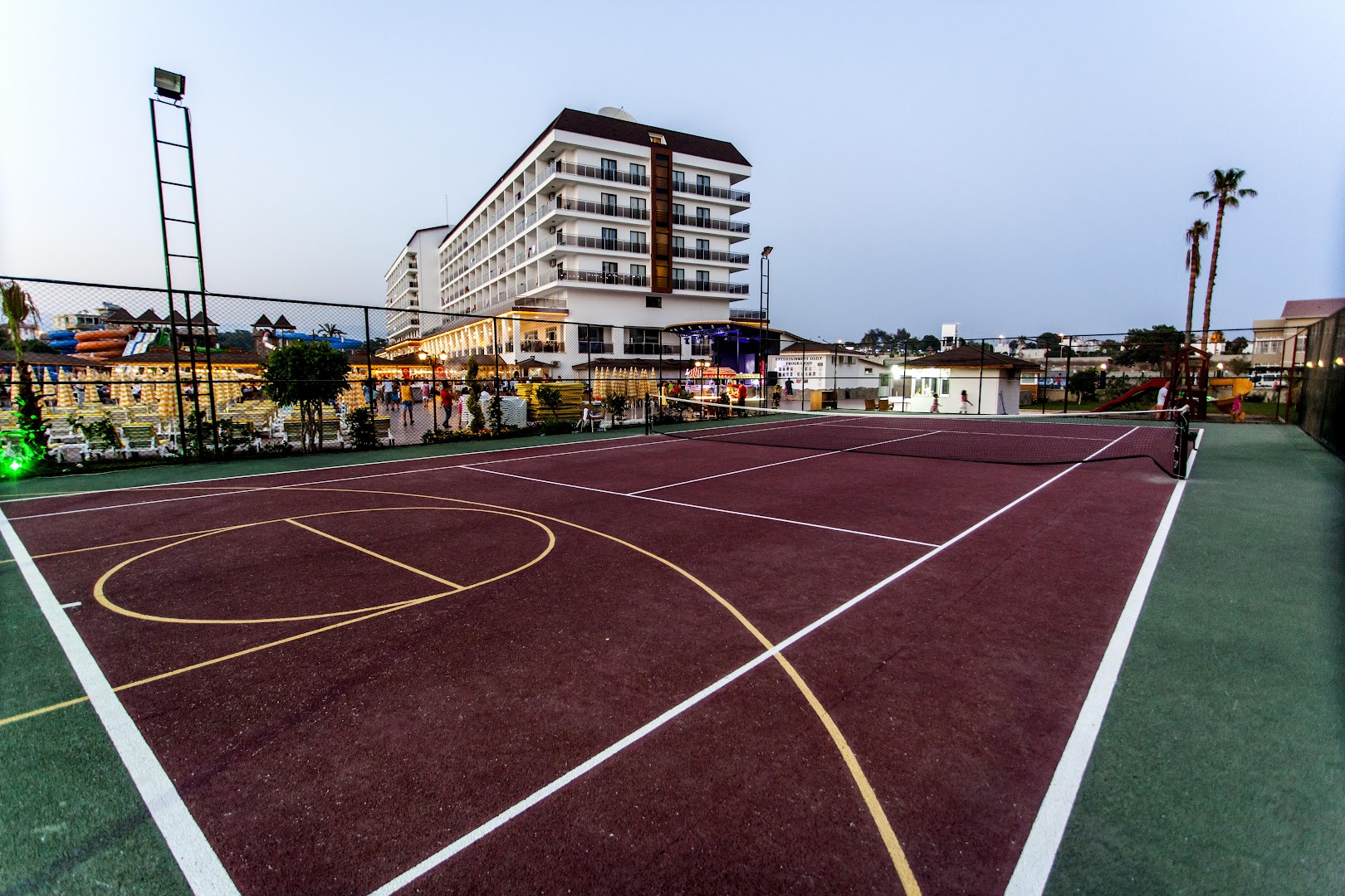 Eftalia Splash Resort daudzfunkcionālais sporta laukums tenisam un basketbolam uz viesnīcas ēkas un palmu fona vakara gaismā