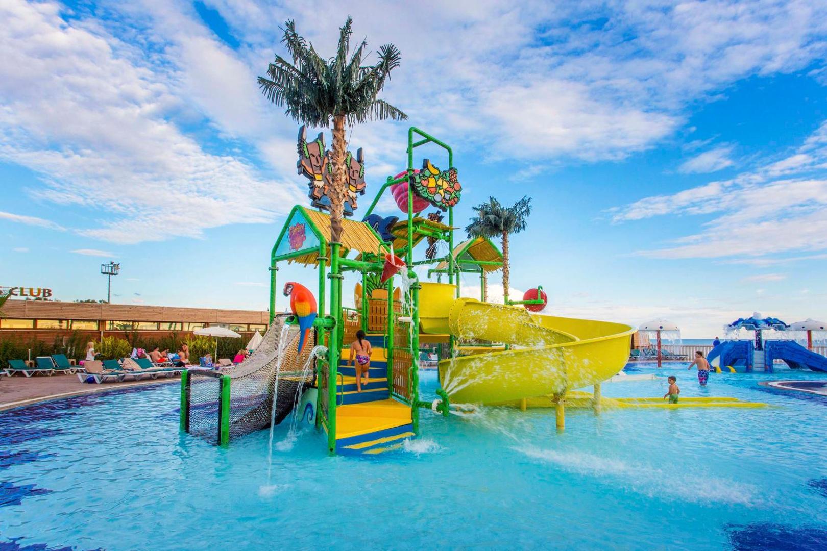 Eftalia Splash Resort bērnu ūdens atrakciju parks ar dzeltenu slidkalniņu, palmu dekorācijām un seklu baseinu saulainā dienā