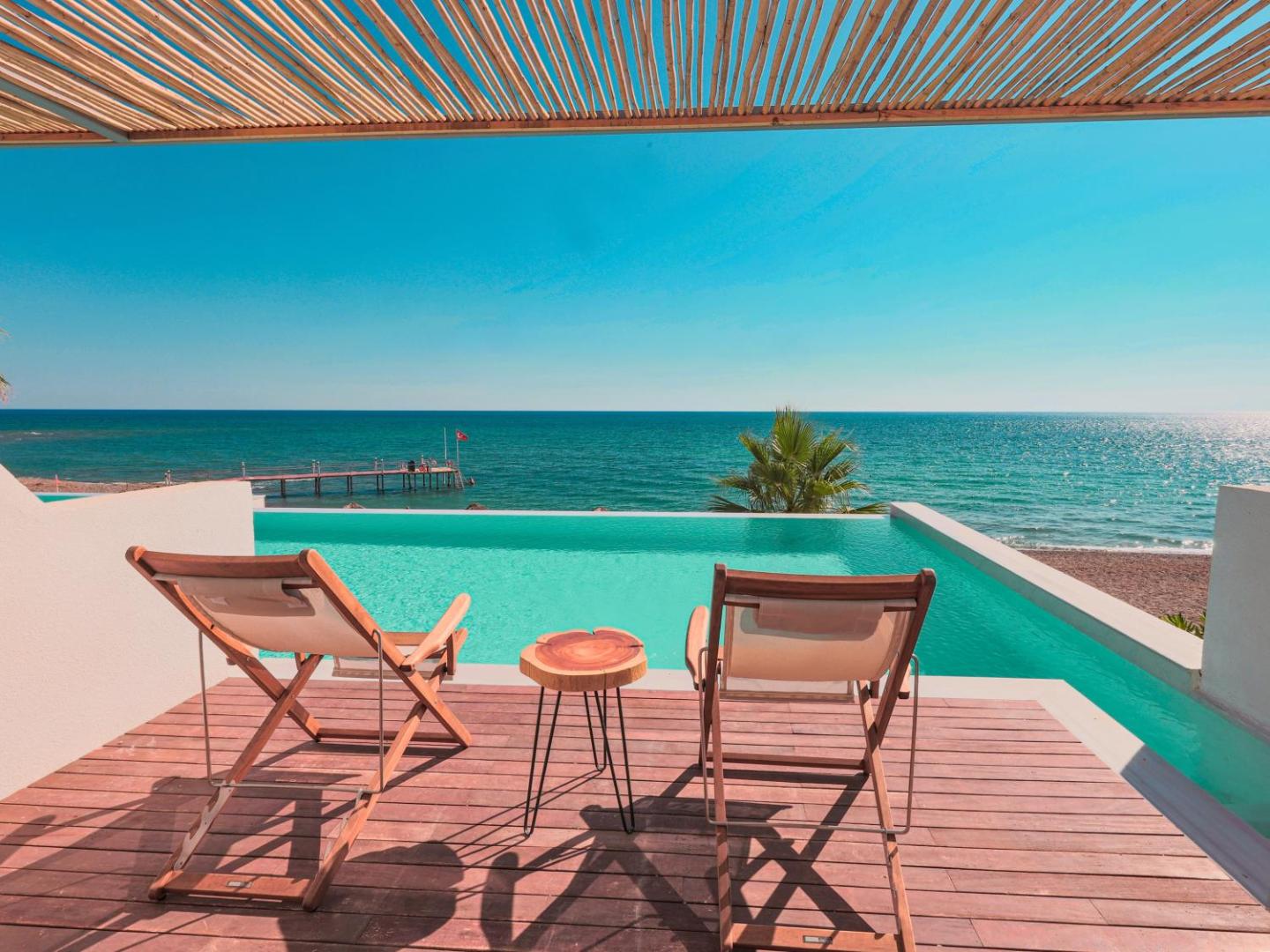 Flora Garden Beach skats no terases uz bezgalības baseinu un Vidusjūru, koka klājs ar sauļošanās krēsliem un nelielu galdiņu.