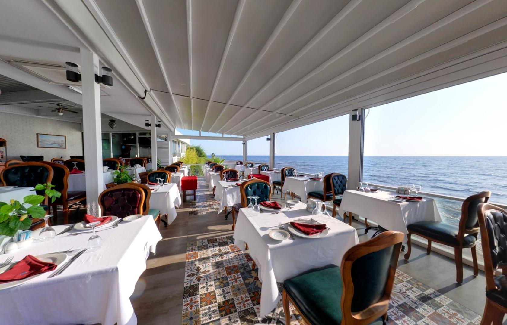 Flora Garden Beach restorāns ar panorāmas logiem un skatu uz Vidusjūru, balti klāti galdi un klasiska stila mēbeles