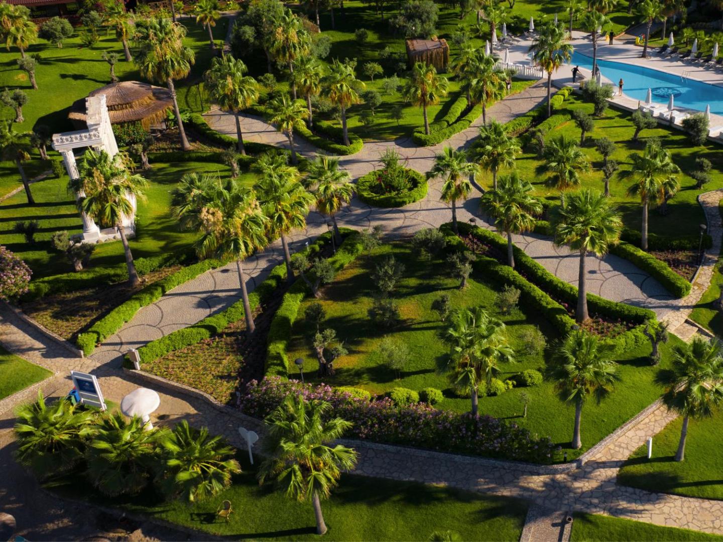 Flora Garden Beach koptā dārza teritorija ar bruģētiem celiņiem, palmām un antīka stila kolonnām saules gaismā