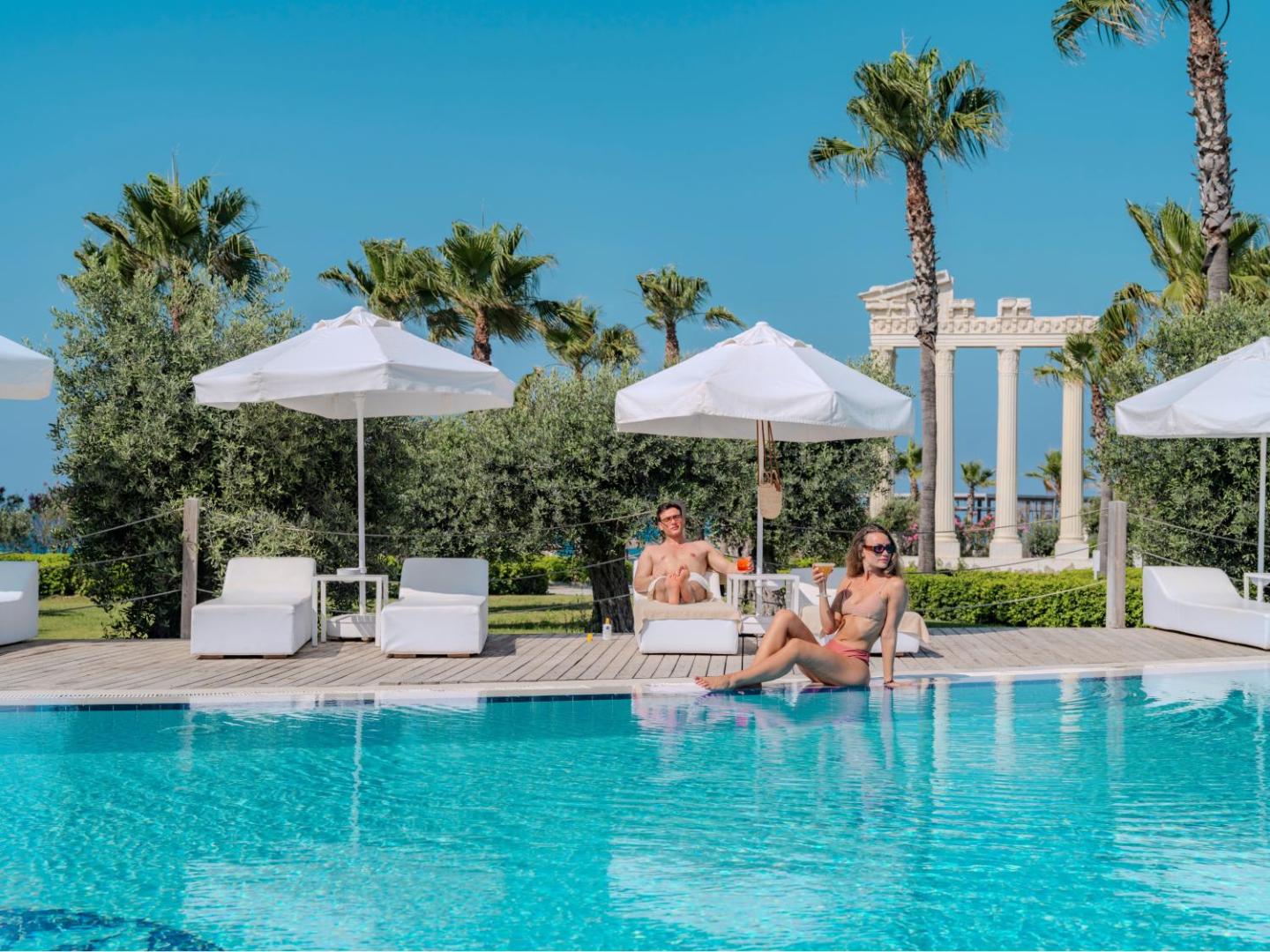 Atpūtnieki pie Flora Garden Beach baseina — balti sauļošanās krēsli, palmas un antīka stila kolonnas fonā