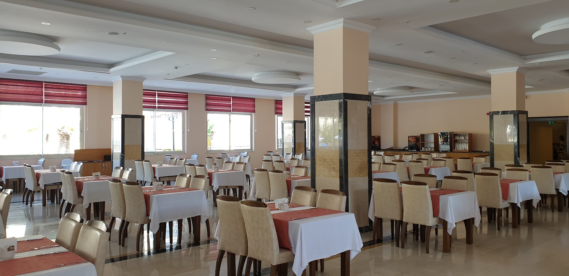 Kemer Dream Hotel galvenais restorāns ar daudziem galdiņiem, baltiem galdautiem un sarkanām detaļām Kemeras reģionā, Turcijā.