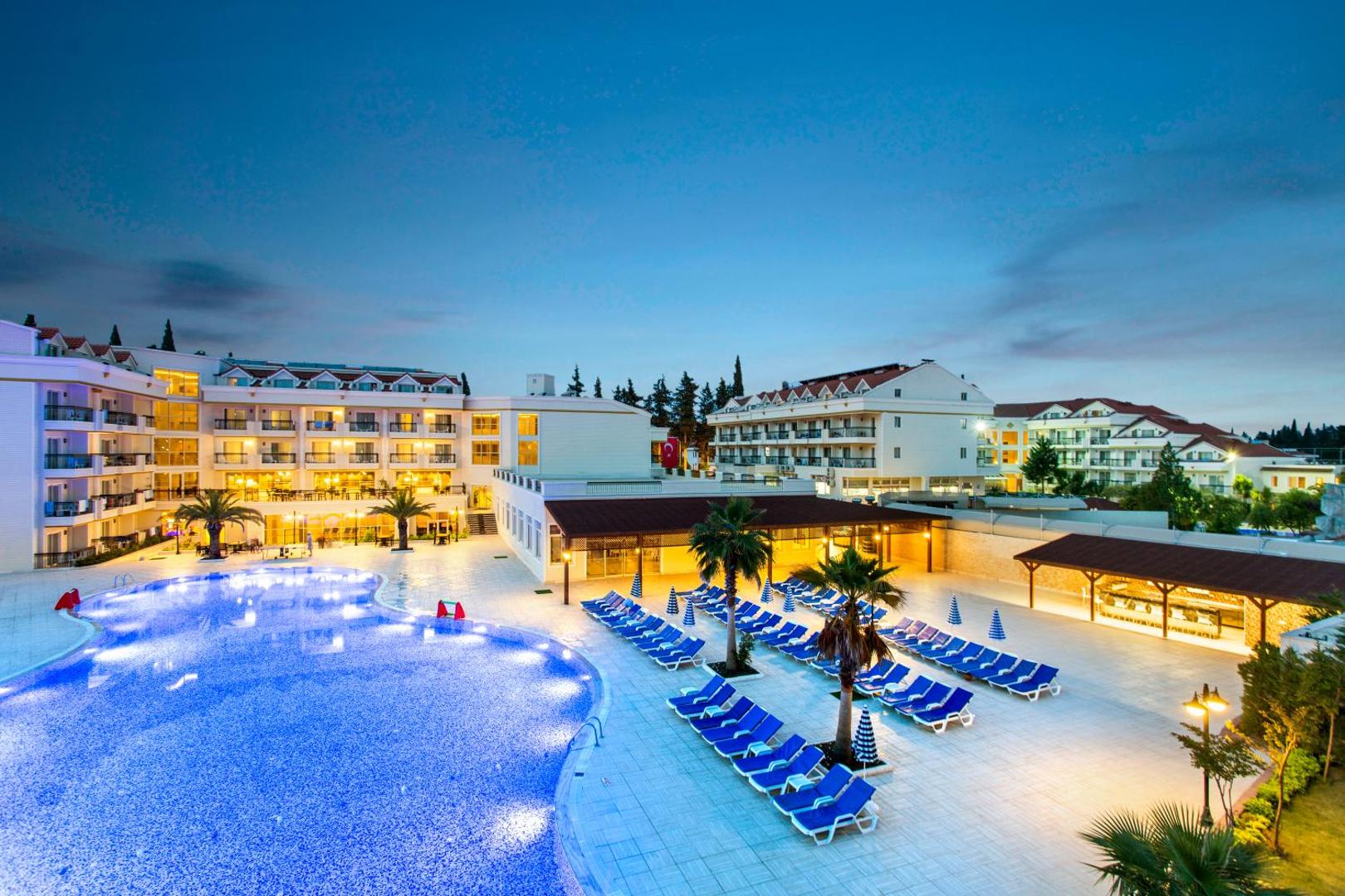 Kemer Dream Hotel plašā atpūtas zona ar baseinu, palmām un ziliem sauļošanās krēsliem vakara apgaismojumā