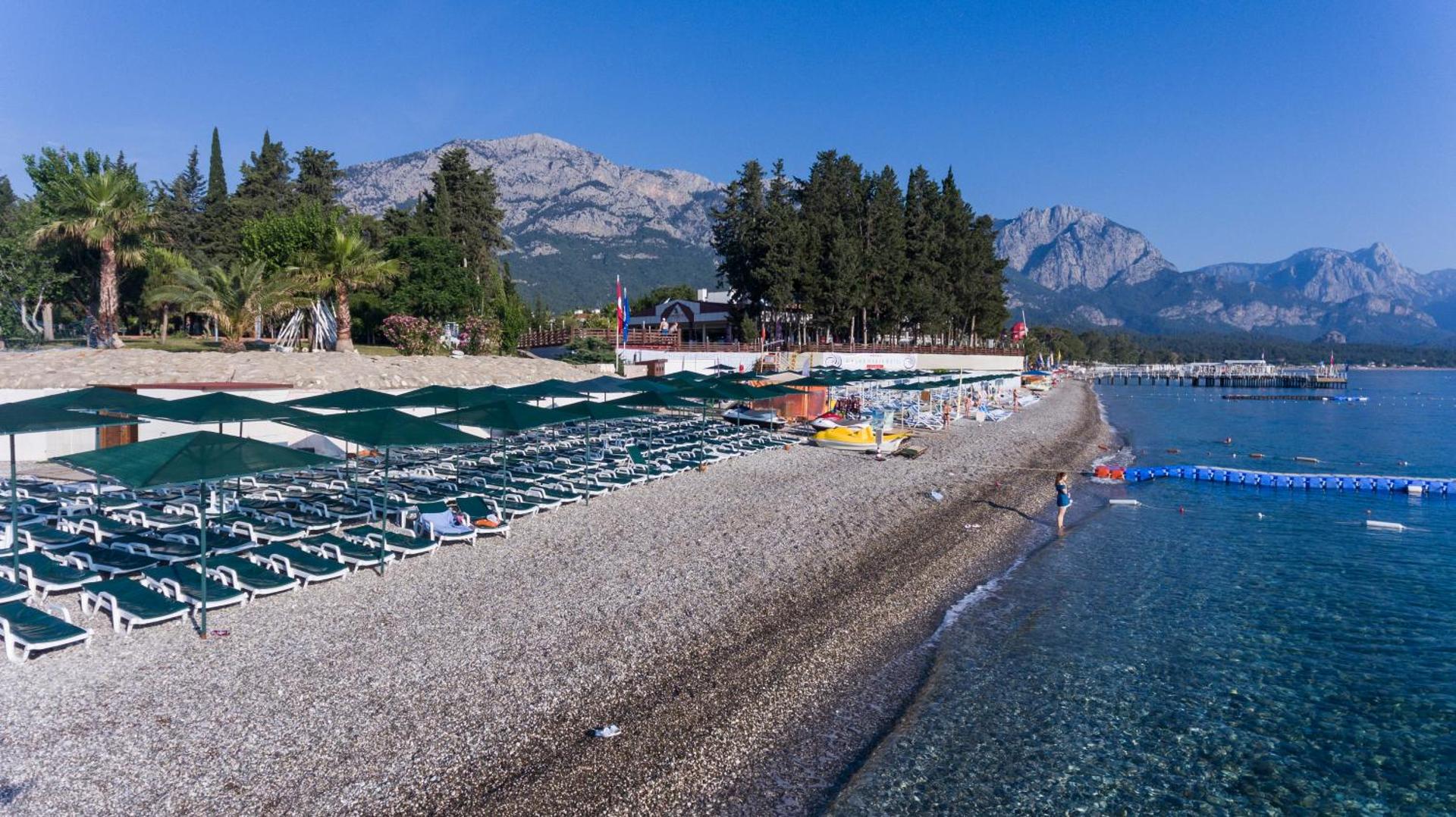 Kemer Dream Hotel oļu pludmale Kemeras piekrastē ar saulessargiem, sauļošanās krēsliem un skatu uz kalniem un dzidru jūras ūdeni