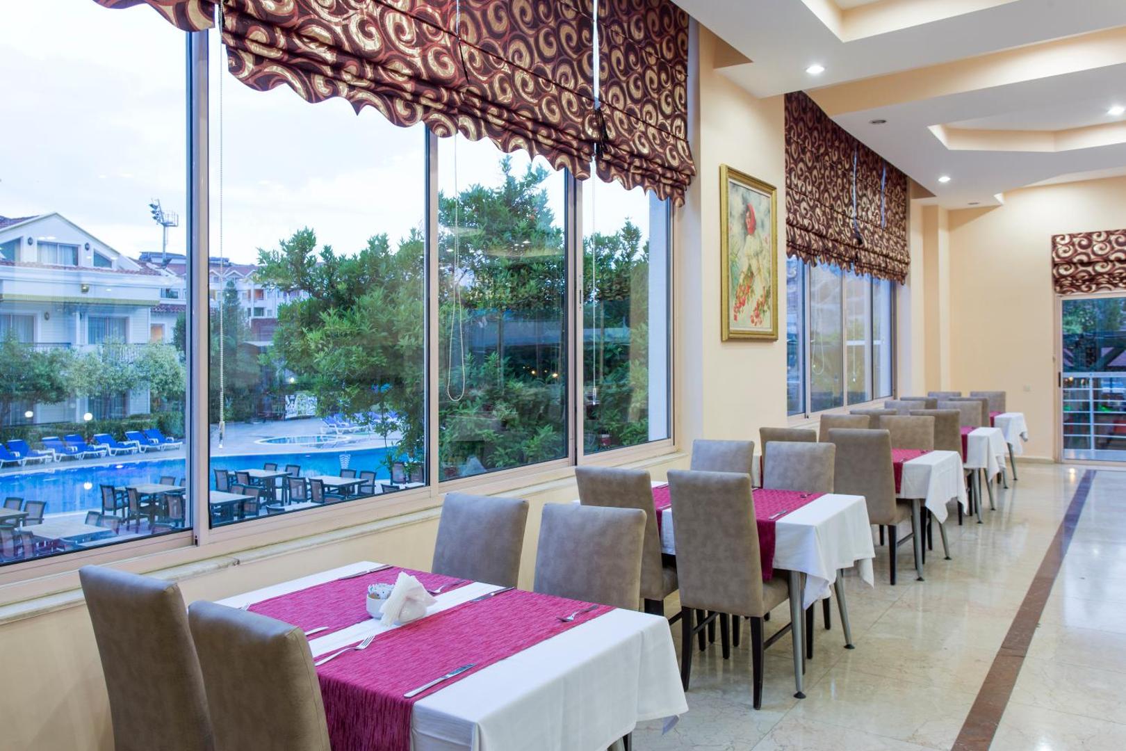 Restorāna zona Kemer Dream Hotel viesnīcā ar lieliem logiem, no kuriem paveras skats uz āra baseinu un zaļo teritoriju