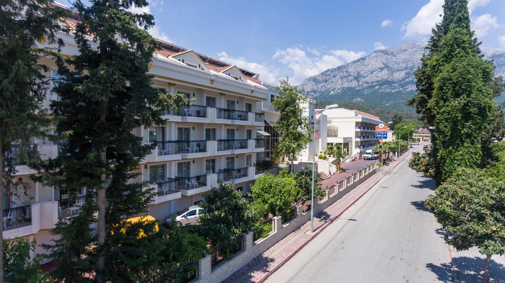 Kemer Dream Hotel ēkas fasāde ar balkoniem un skatu uz ielu, fonā redzami augsti kalni un zaļojoši koki.