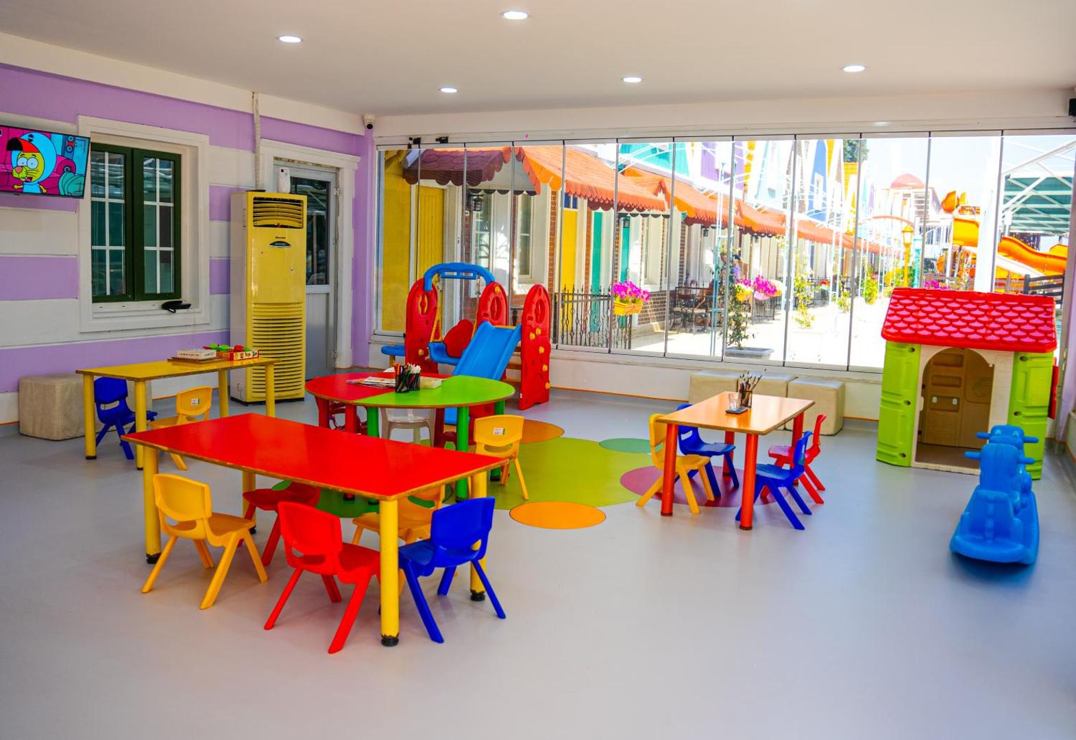 Orange County Kemer bērnu kluba iekštelpas ar krāsainiem galdiem, krēsliem un rotaļlietām — gaiša rotaļu zona ar skatu uz pagalmu