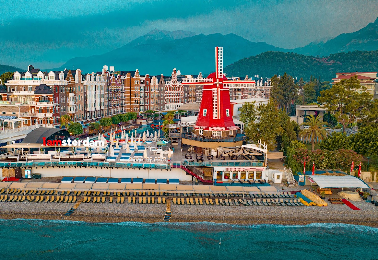 Orange County Kemer viesnīcas panorāmas skats no jūras puses — pludmale, Amsterdamas stila arhitektūra un sarkanas vējdzirnavas Kemeras piekrastē