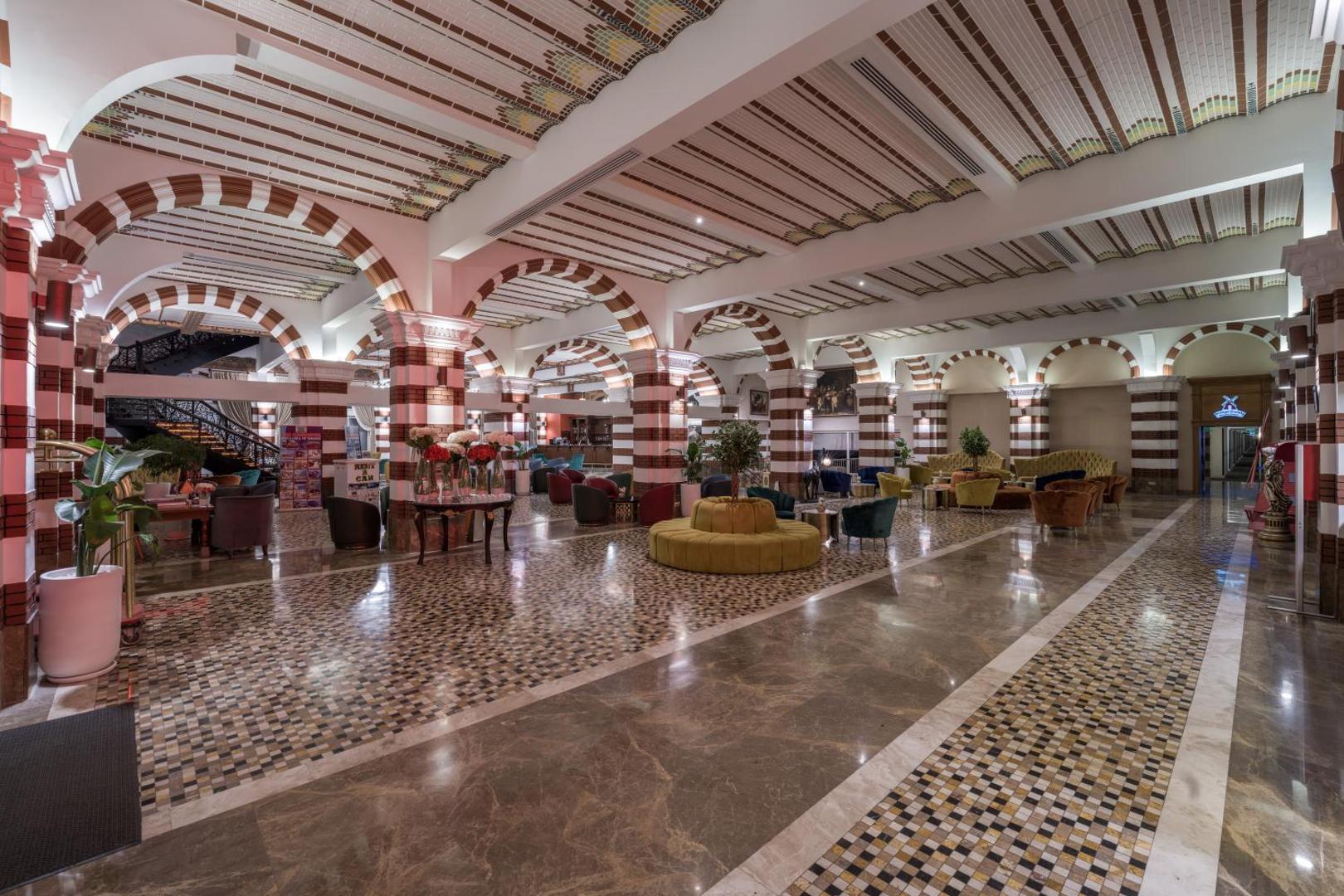 Orange County Kemer viesnīcas plašais lobby bārs un atpūtas zona ar arkām, mozaīkas grīdu un krāsainiem dīvāniem
