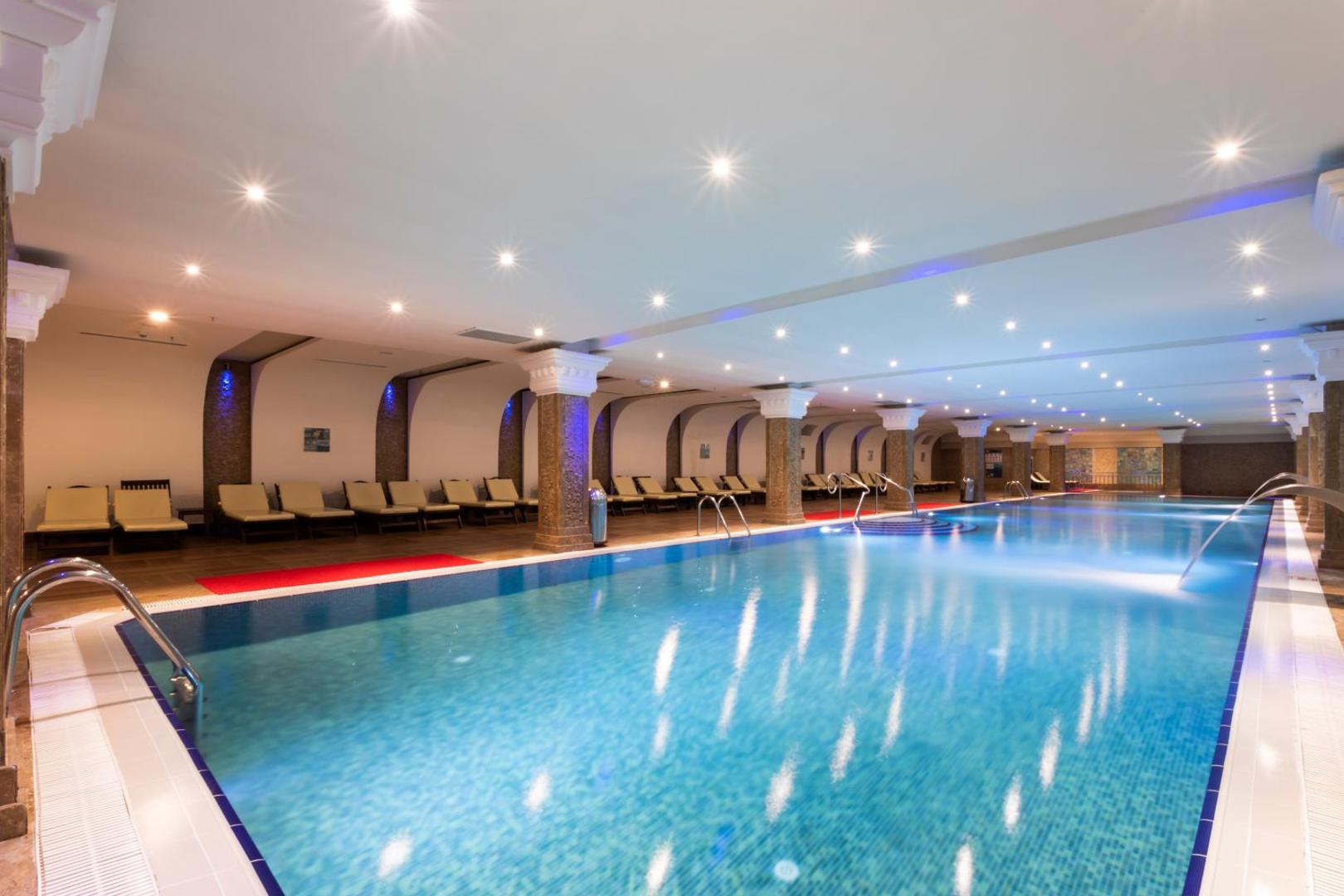 Orange County Kemer iekštelpu baseins ar apsildi un sauļošanās krēsliem — spa zona ar elegantām kolonnām un mierīgu apgaismojumu