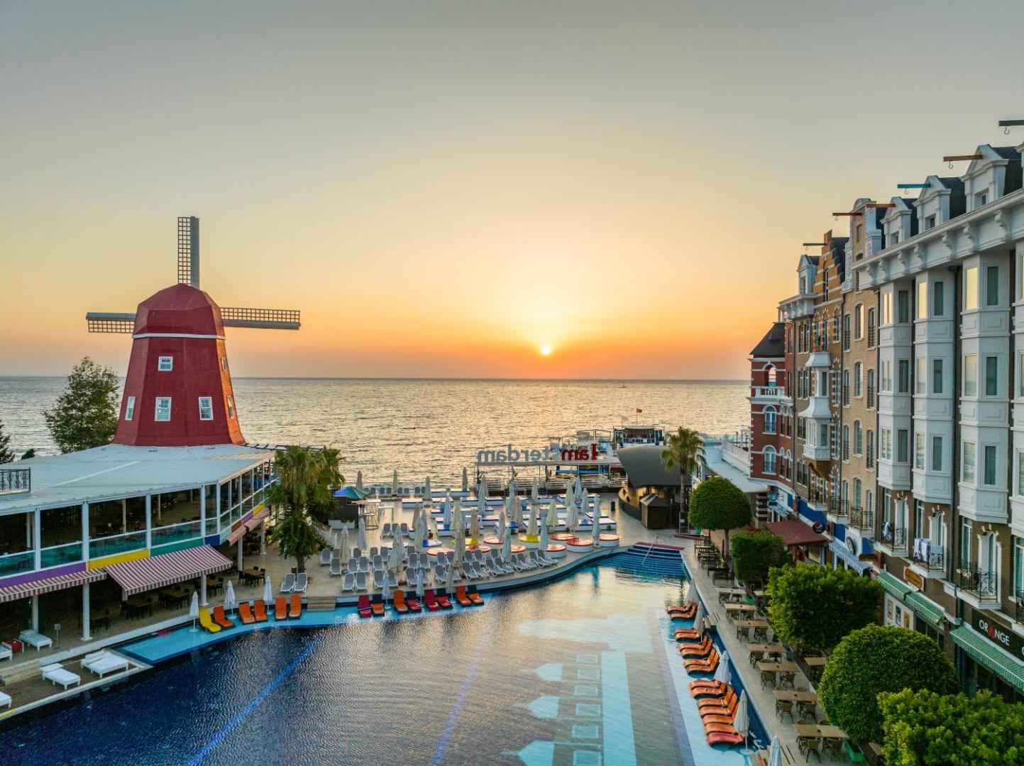 Orange County Kemer viesnīcas teritorija saulrietā ar baseinu, dekoratīvām vējdzirnavām un Amsterdamas stila arhitektūru Kemeras piekrastē