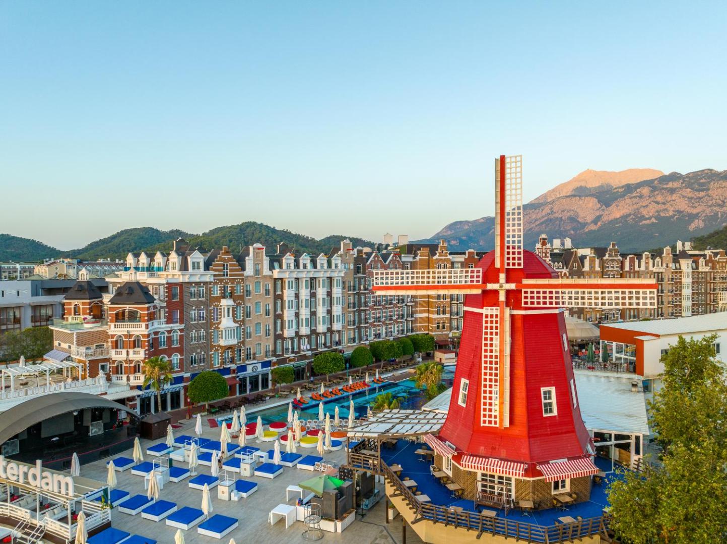 Orange County Kemer centrālā zona ar ikoniskajām sarkanajām vējdzirnavām, sauļošanās krēsliem un Amsterdamas stila ēkām uz kalnu fona