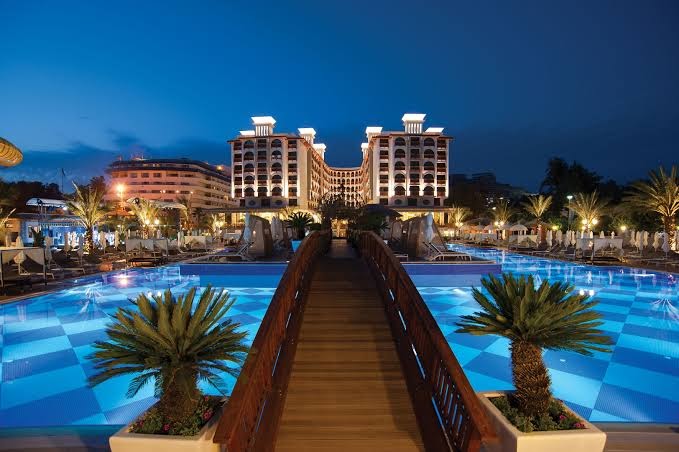 Quattro Beach Spa & Resort vakara skats uz izgaismotu baseinu un viesnīcas ēku ar koka tiltiņu un palmām