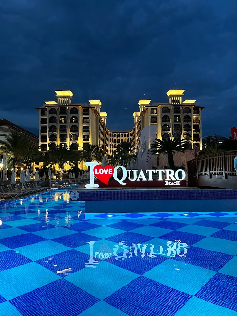 Quattro Beach Spa & Resort apgaismots āra baseins vakarā ar viesnīcas fasādi un palmu siluetiem Alānijas reģionā, Turcijā