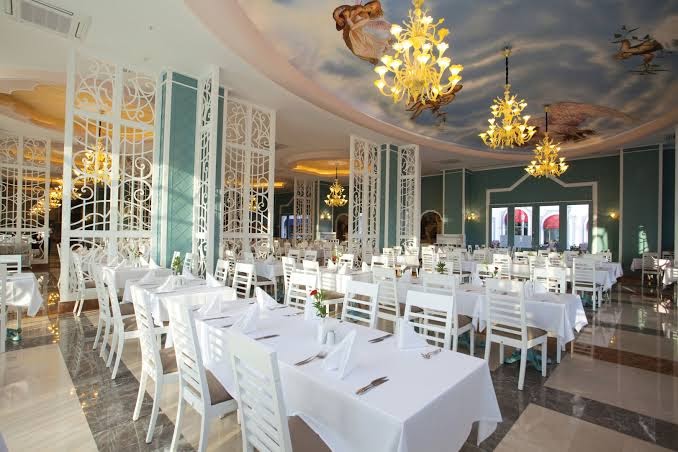 Quattro Beach Spa & Resort eleganta restorāna zāle ar greznām lustrām, apgleznotiem griestiem un balti klātiem galdiem