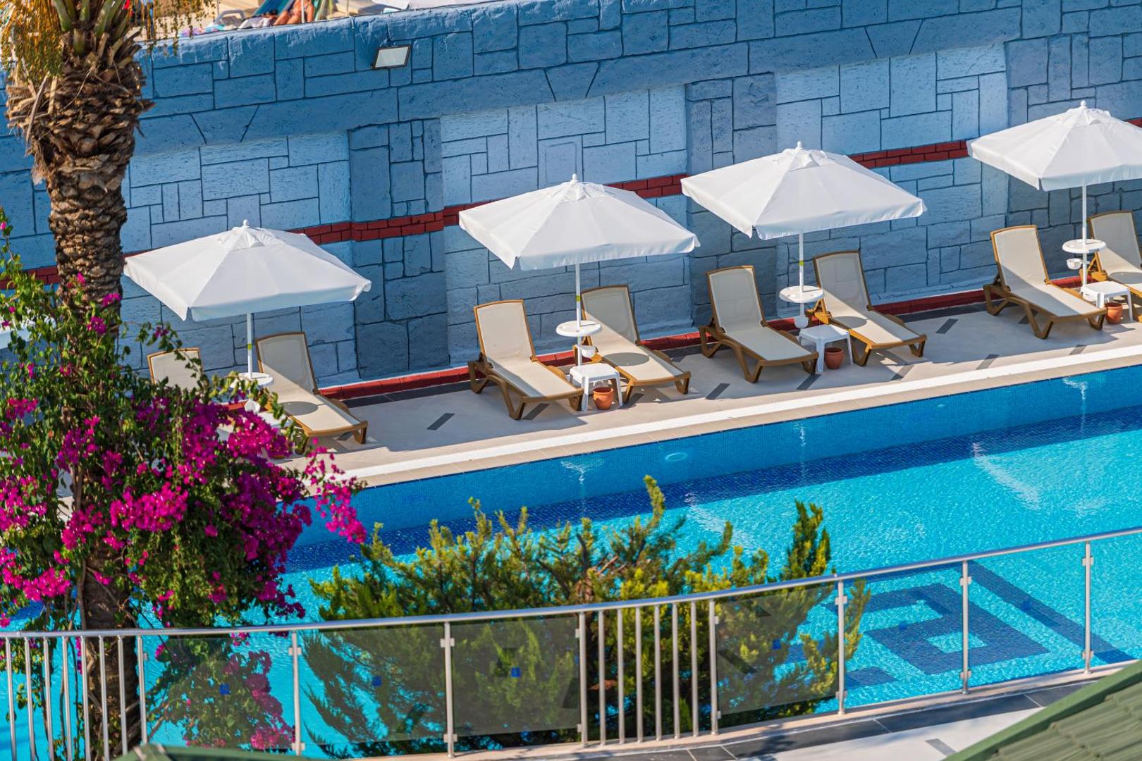 Sealife Kemer Resort Hotel sauļošanās zona pie baseina ar ērtiem zviļņiem, baltiem saulessargiem un ziedošiem krūmiem