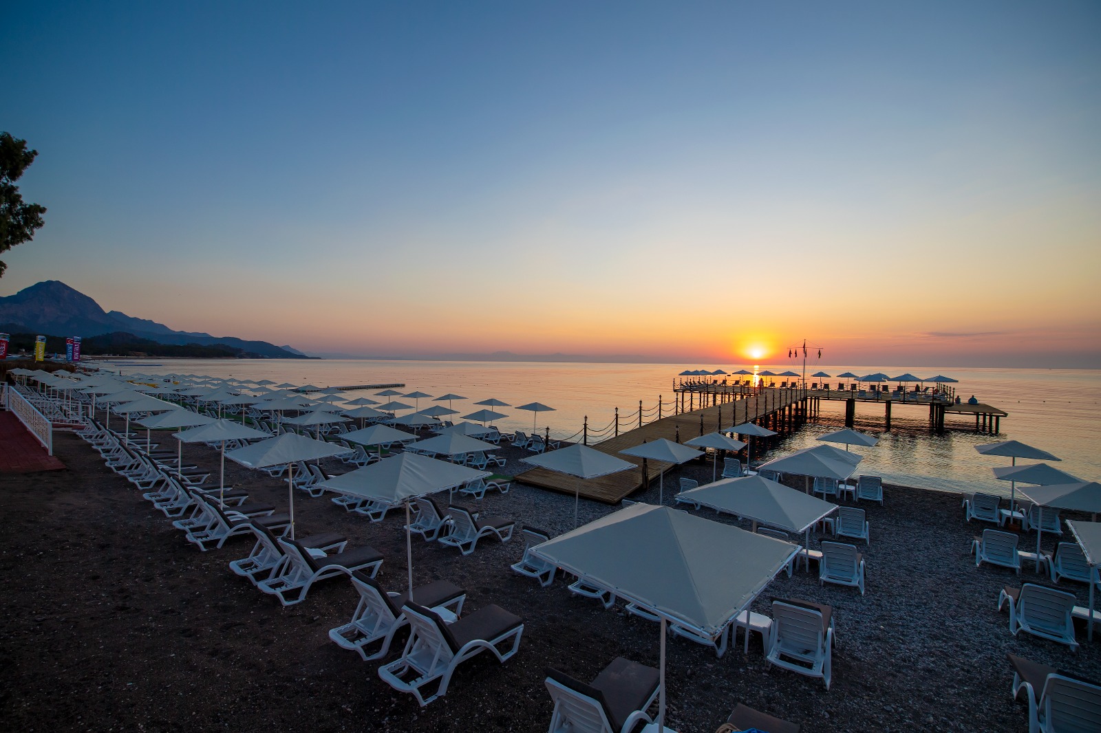 Sealife Kemer Resort Hotel pludmales zona un koka mols saulrieta laikā, balti saulessargi un mierīga jūra