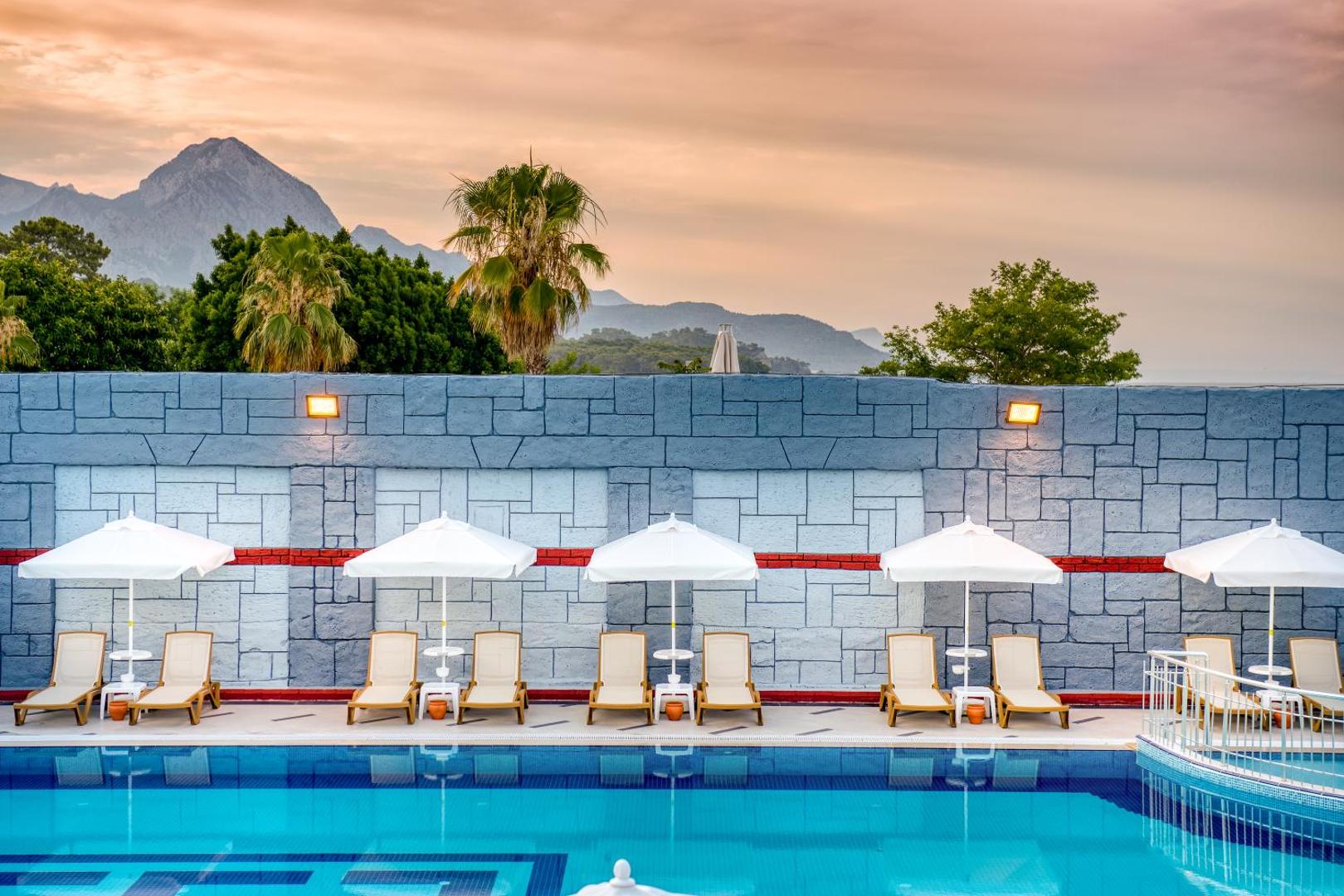 Sealife Kemer Resort Hotel baseina zona ar baltiem saulessargiem un zviļņiem uz iespaidīgu kalnu fona saulrietā