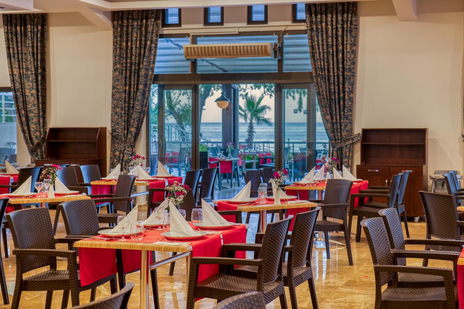 Sealife Kemer Resort Hotel iekštelpu restorāns ar eleganti klātiem galdiem, sarkaniem galdautiem un skatu caur logu uz palmu dārzu