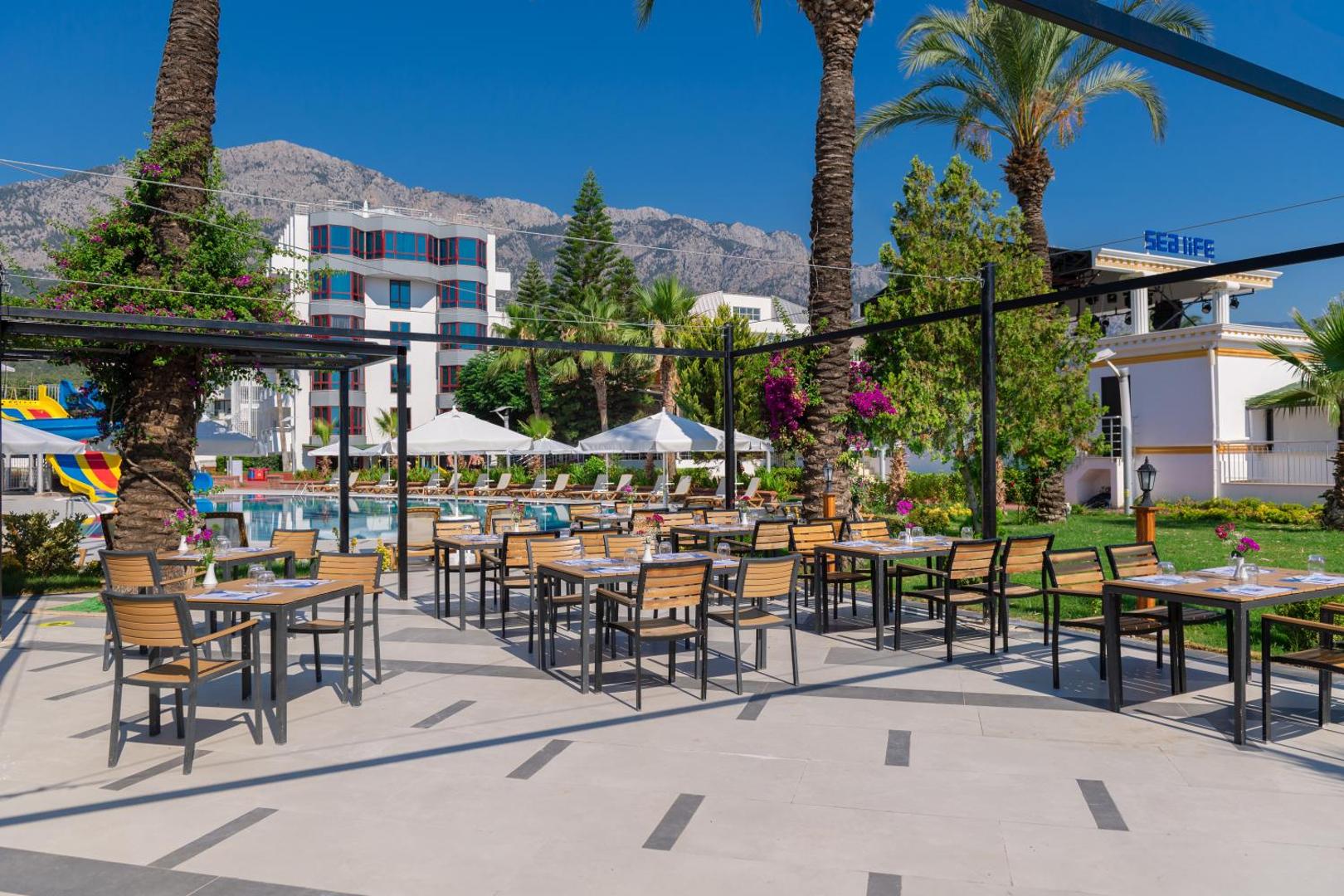 Sealife Kemer Resort Hotel āra restorāna zona ar skatu uz viesnīcas korpusu, baseinu un iespaidīgajiem Kemeras kalniem