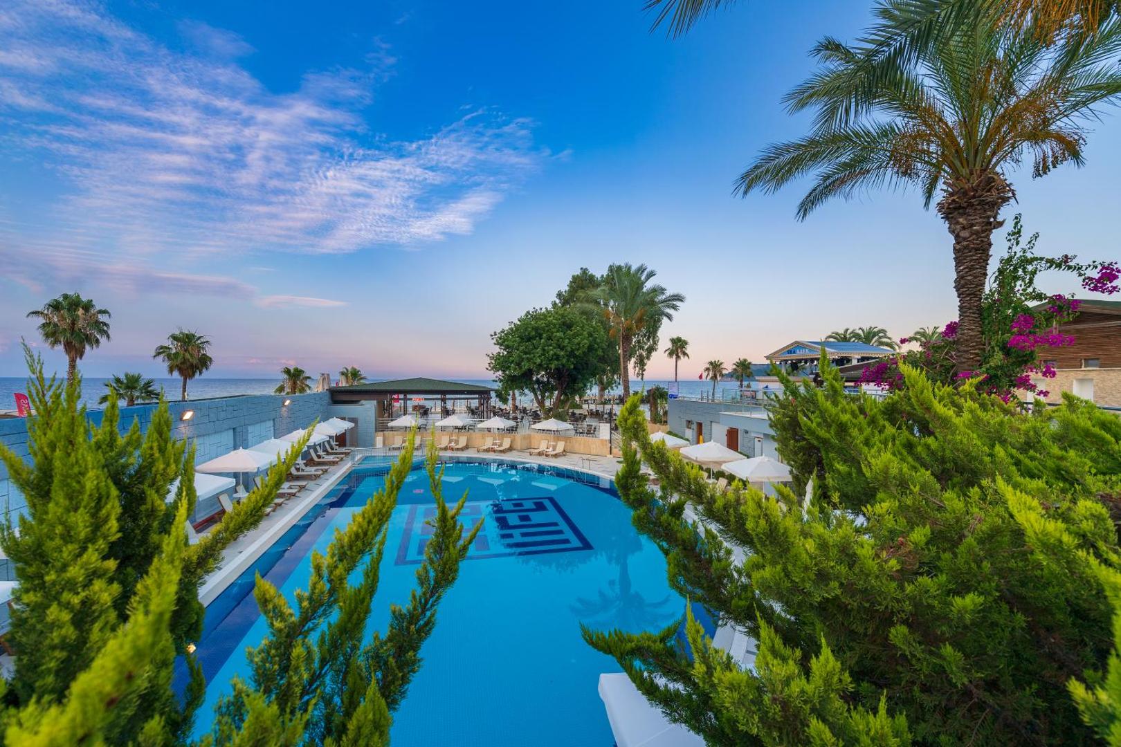 Sealife Kemer Resort Hotel plašais āra baseins, ko ieskauj palmas un zaļumi, ar skatu uz jūru un kalniem tālumā