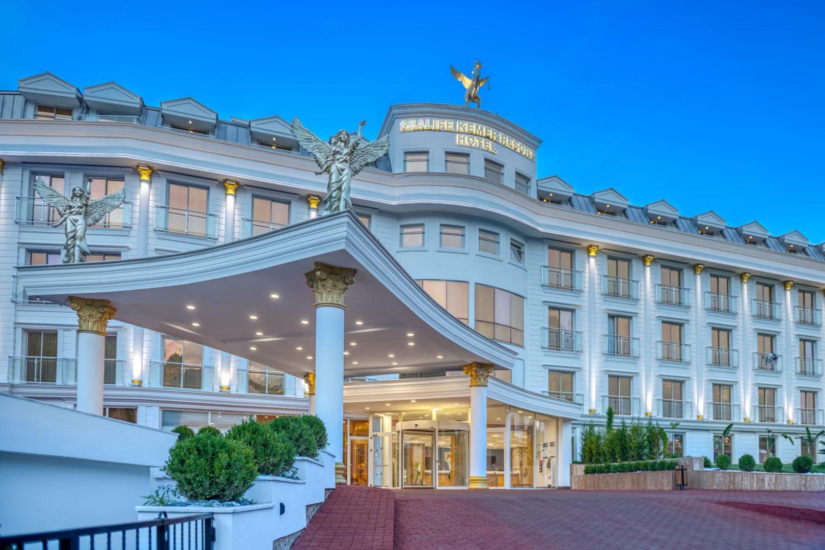 Sealife Kemer Resort galvenā ieeja ar klasiskām kolonnām un eņģeļu skulptūrām vakara apgaismojumā Kemerā, Turcijā