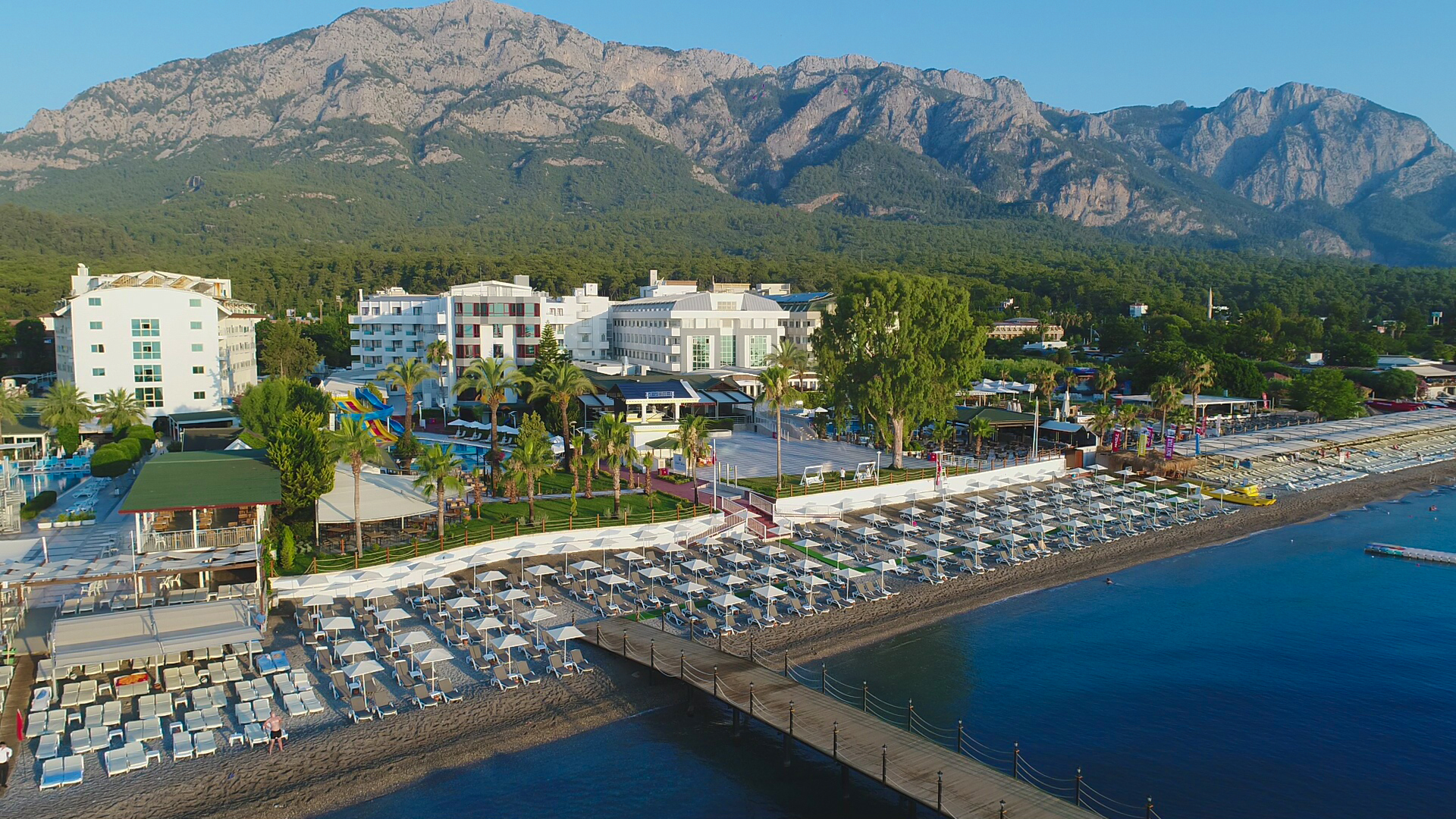 Sealife Kemer Resort skats no putna lidojuma uz pludmali, piestātni un viesnīcas ēkām uz majestātisku kalnu fona Kemerā