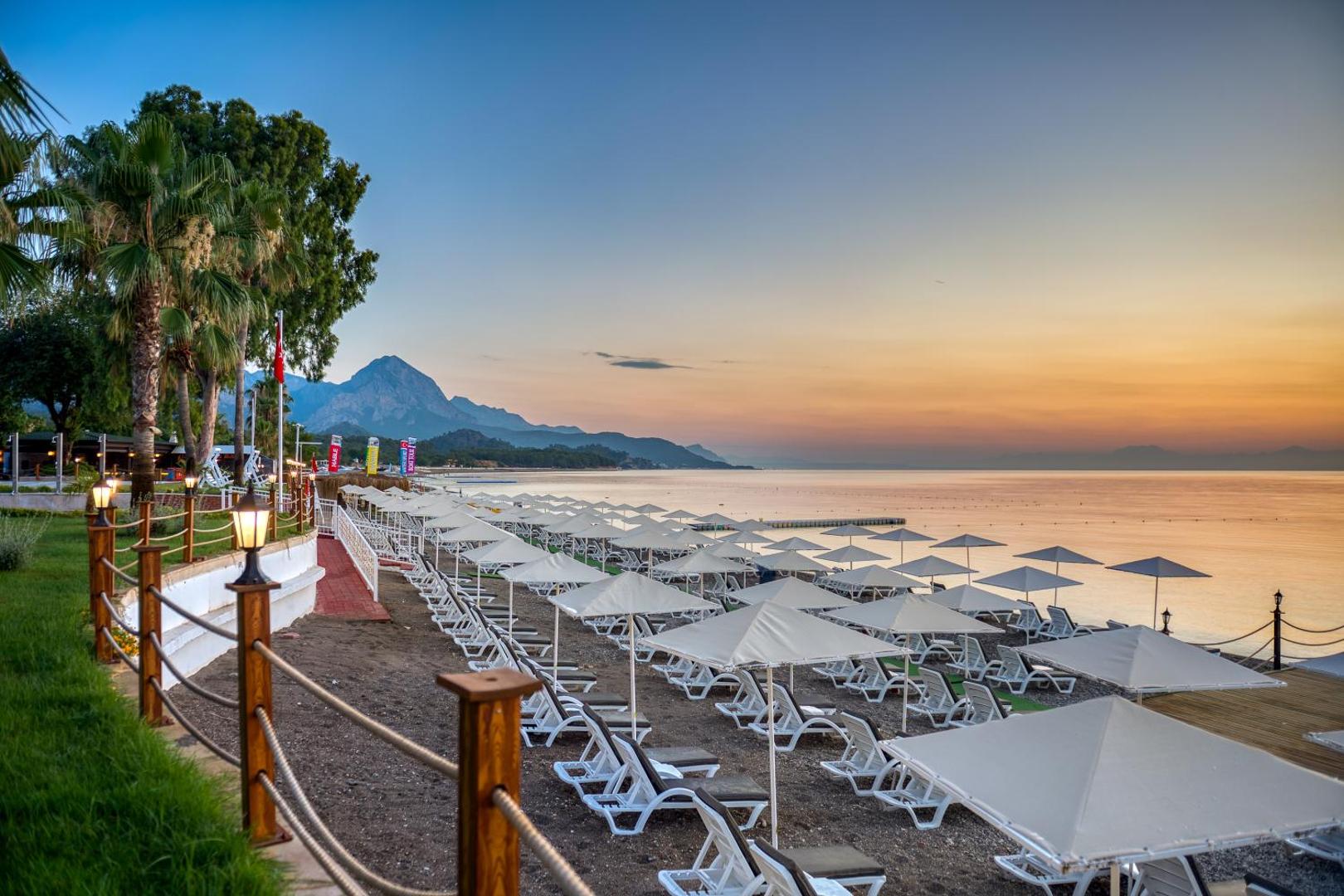 Sealife Kemer Resort Hotel privātā pludmale saulrietā ar baltiem saulessargiem, sauļošanās krēsliem un kalnu fonu Kemerā