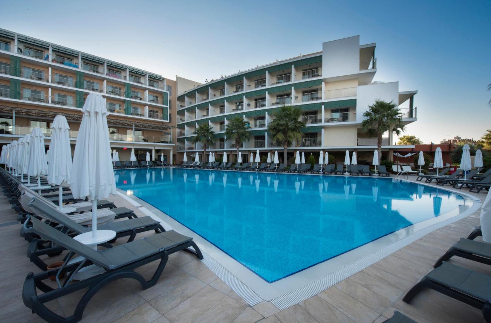 TUI BLUE Barut Andiz plašais āra baseins ar dzidru ūdeni, sauļošanās krēsliem un viesnīcas korpusu fonā dienas gaismā