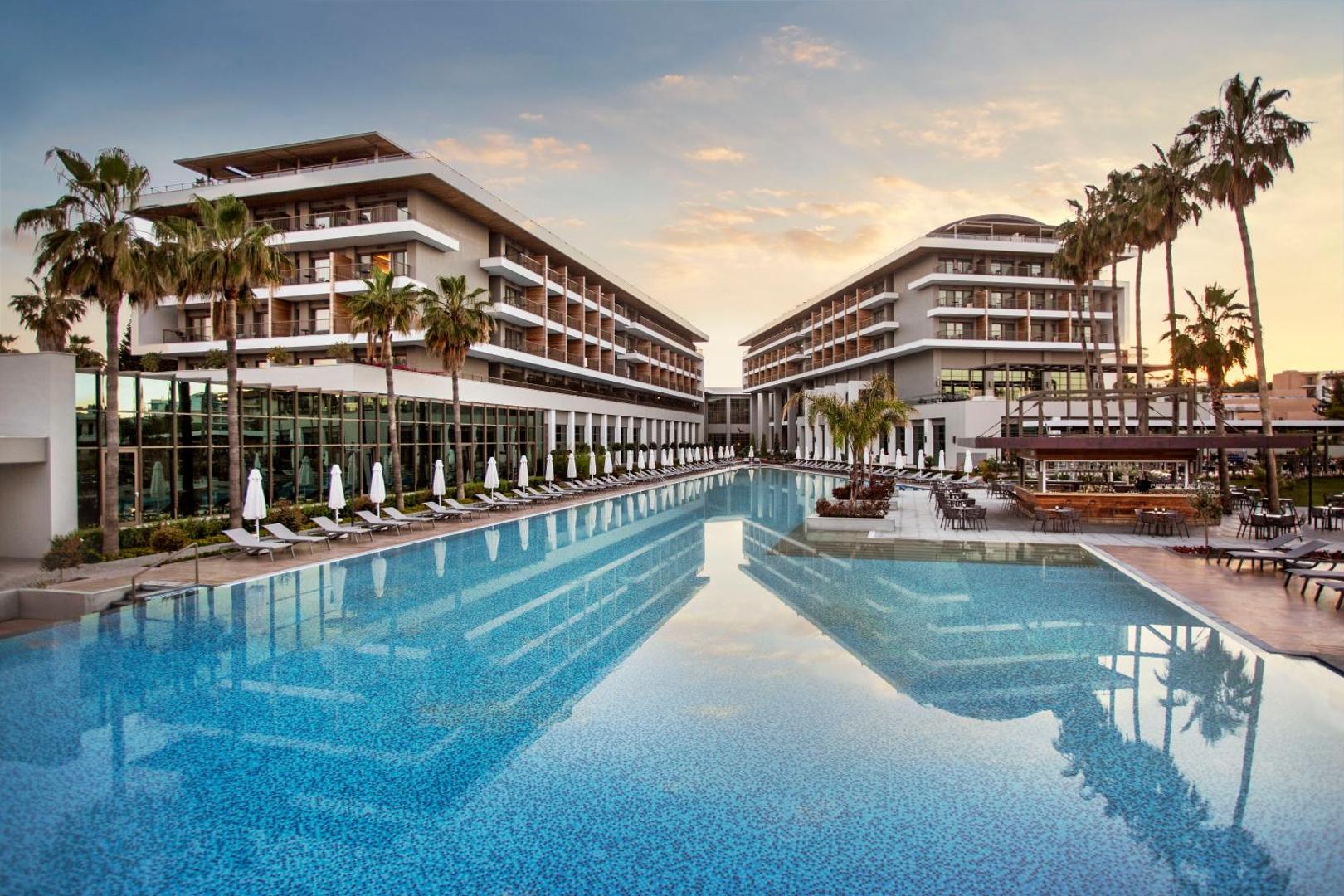 Acanthus & Cennet Barut Collection viesnīcas arhitektūra un garais āra baseins saulrietā Sidē, Turcijā — moderni korpusi ar balkoniem un palmām
