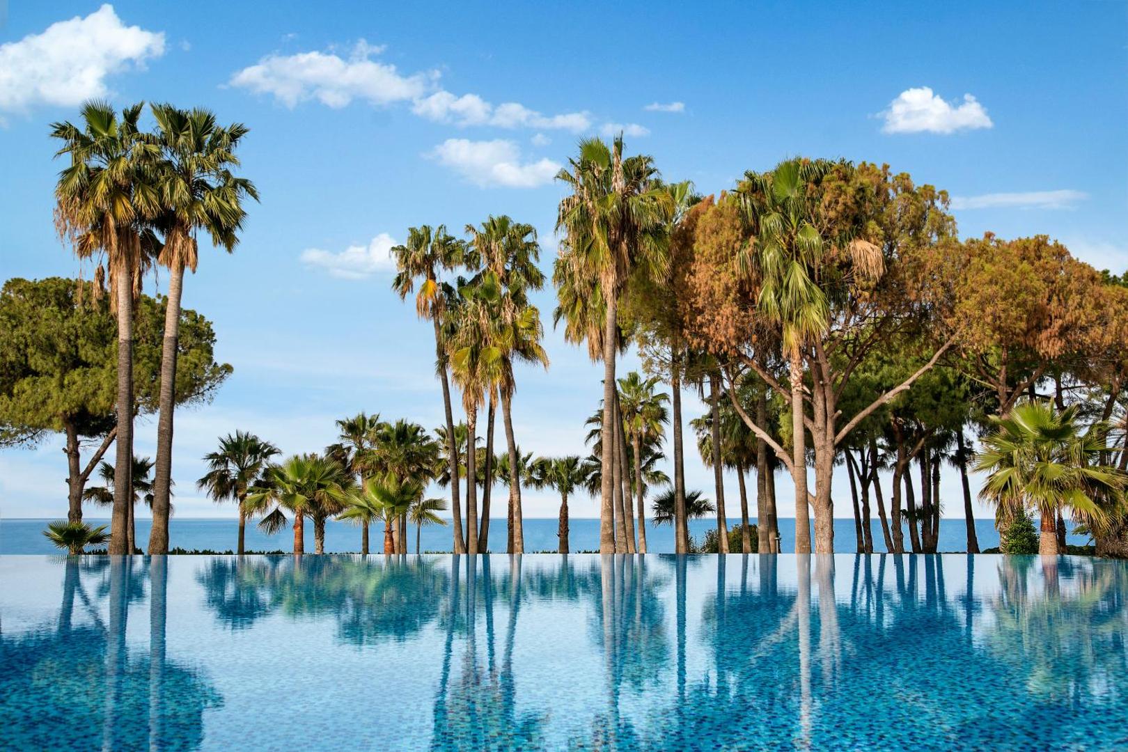 Acanthus & Cennet Barut Collection bezgalības tipa baseina atspulgs ar palmām un Vidusjūras horizontu fonā
