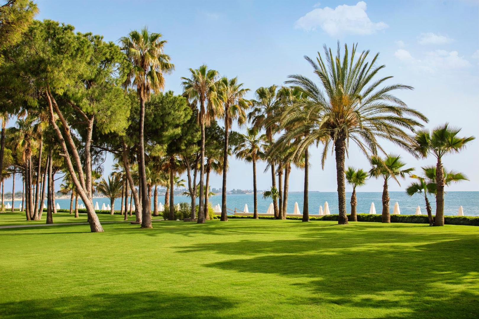 Acanthus & Cennet Barut Collection viesnīcas plašais dārzs ar koptu zālienu un palmām piekrastē Sidē, Turcijā — skats uz jūru